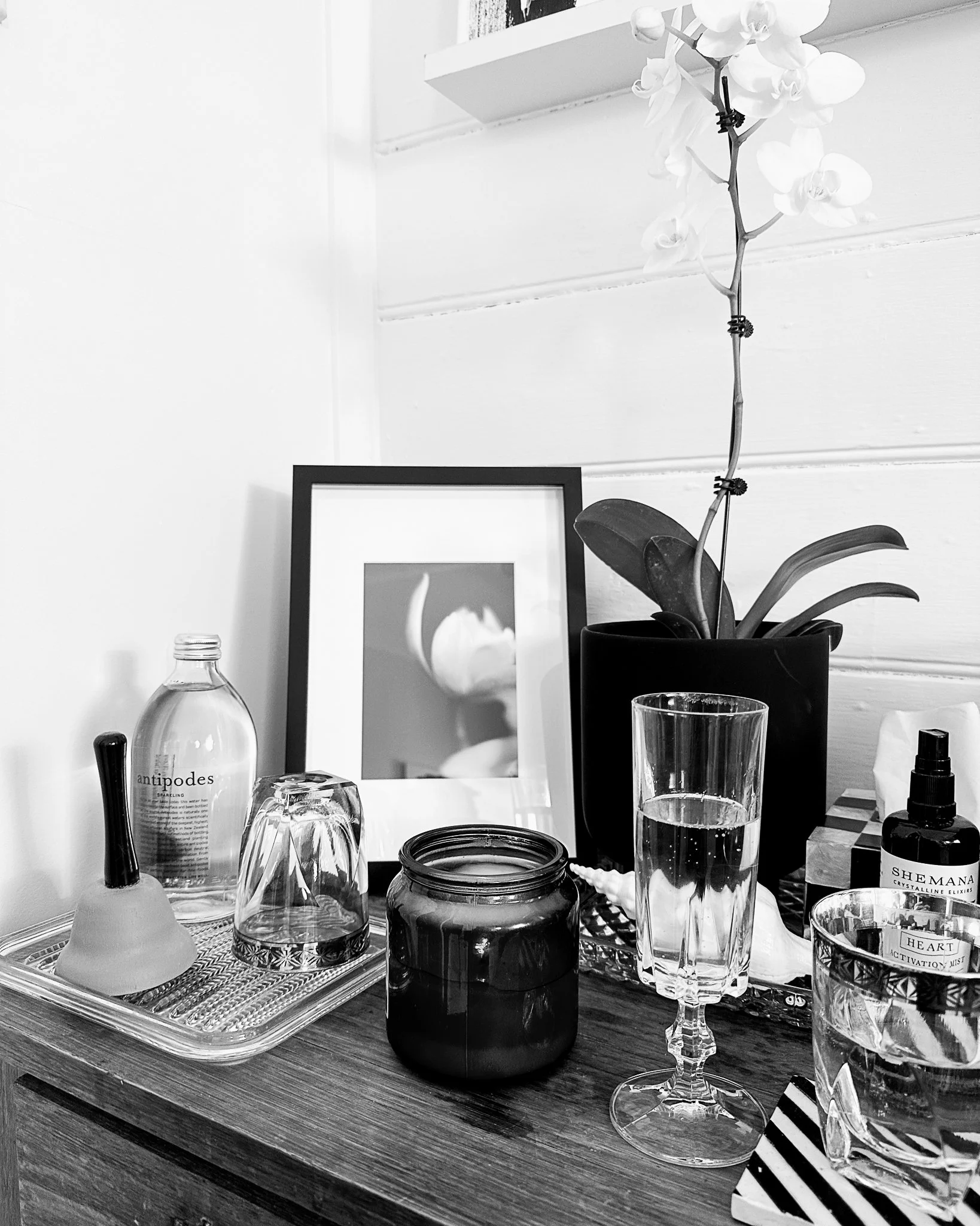 A table with various items including a glass of water, a black candle holder, jars, a small dropper bottle a plate with a striped box, a framed picture leaning against the wall, and an orchid in a black pot. The background shows a white wall with a s