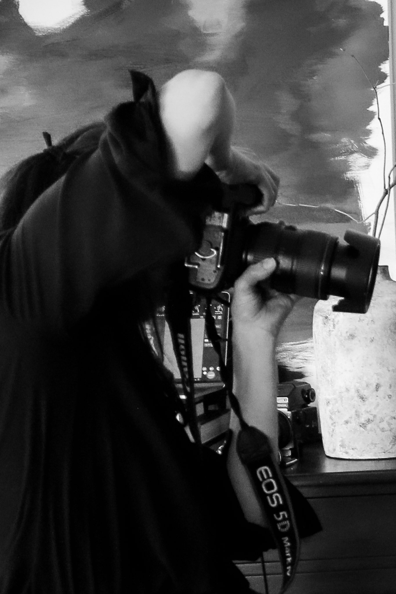 A person with long hair holding a camera, taking a photo of a mirror reflection. The person is wearing a dark jacket and is in a room with various objects, including a white textured vase and electronic equipment in the background.
