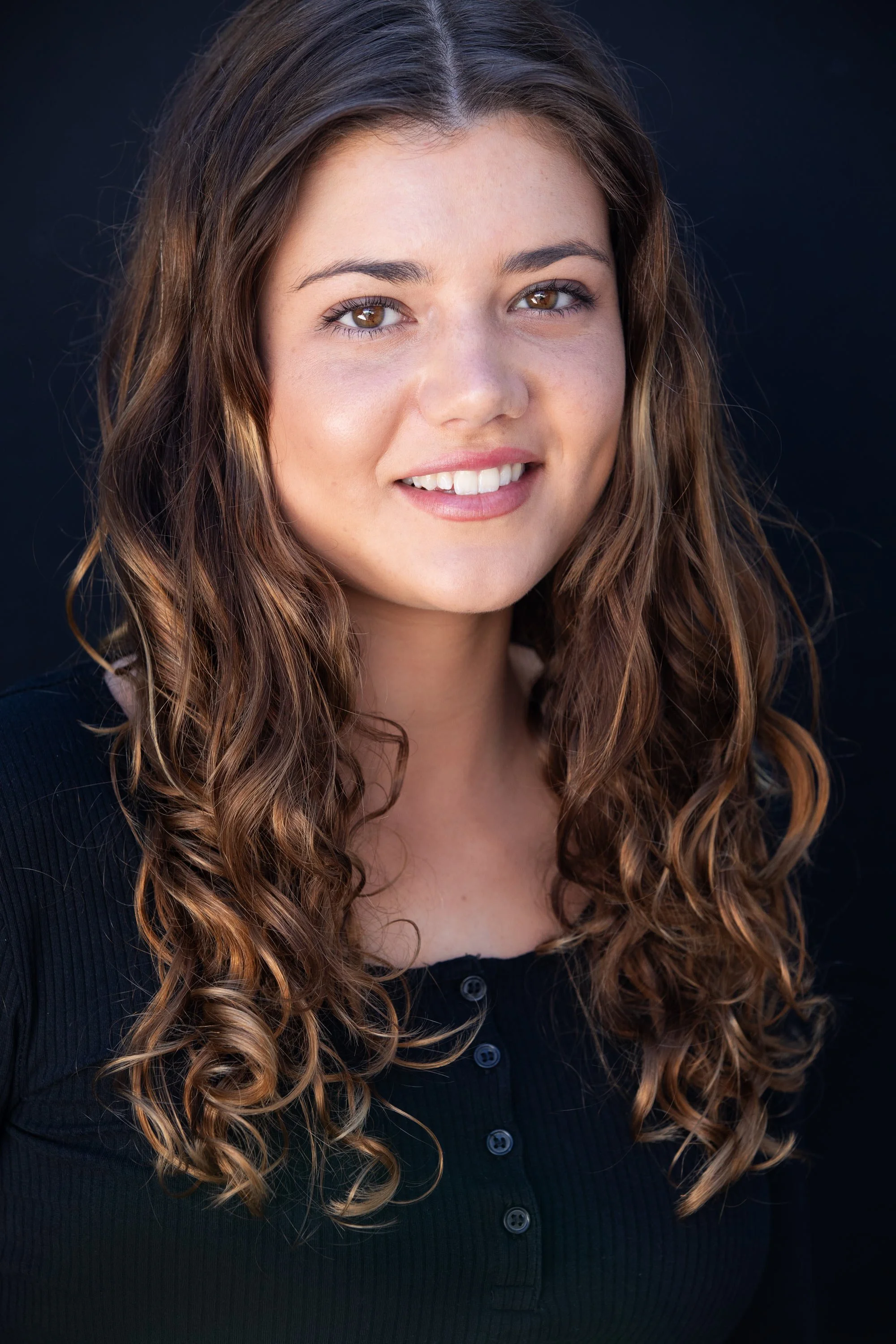 A young woman with long, curly brown hair and light brown eyes, smiling slightly, wearing a black button-up top.