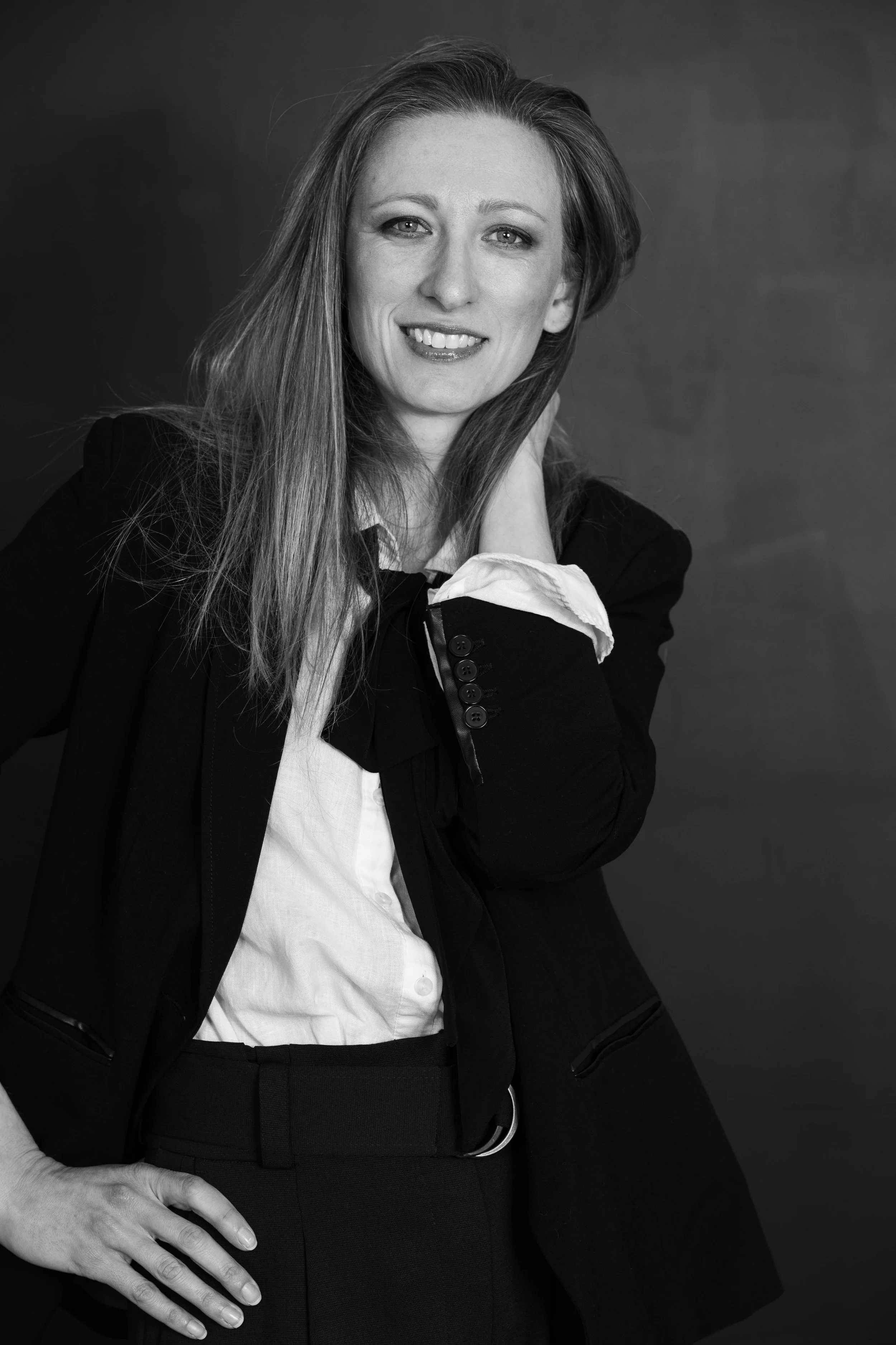 Black and white portrait of a woman with long hair, smiling, wearing a blazer and white shirt, standing against a dark background.