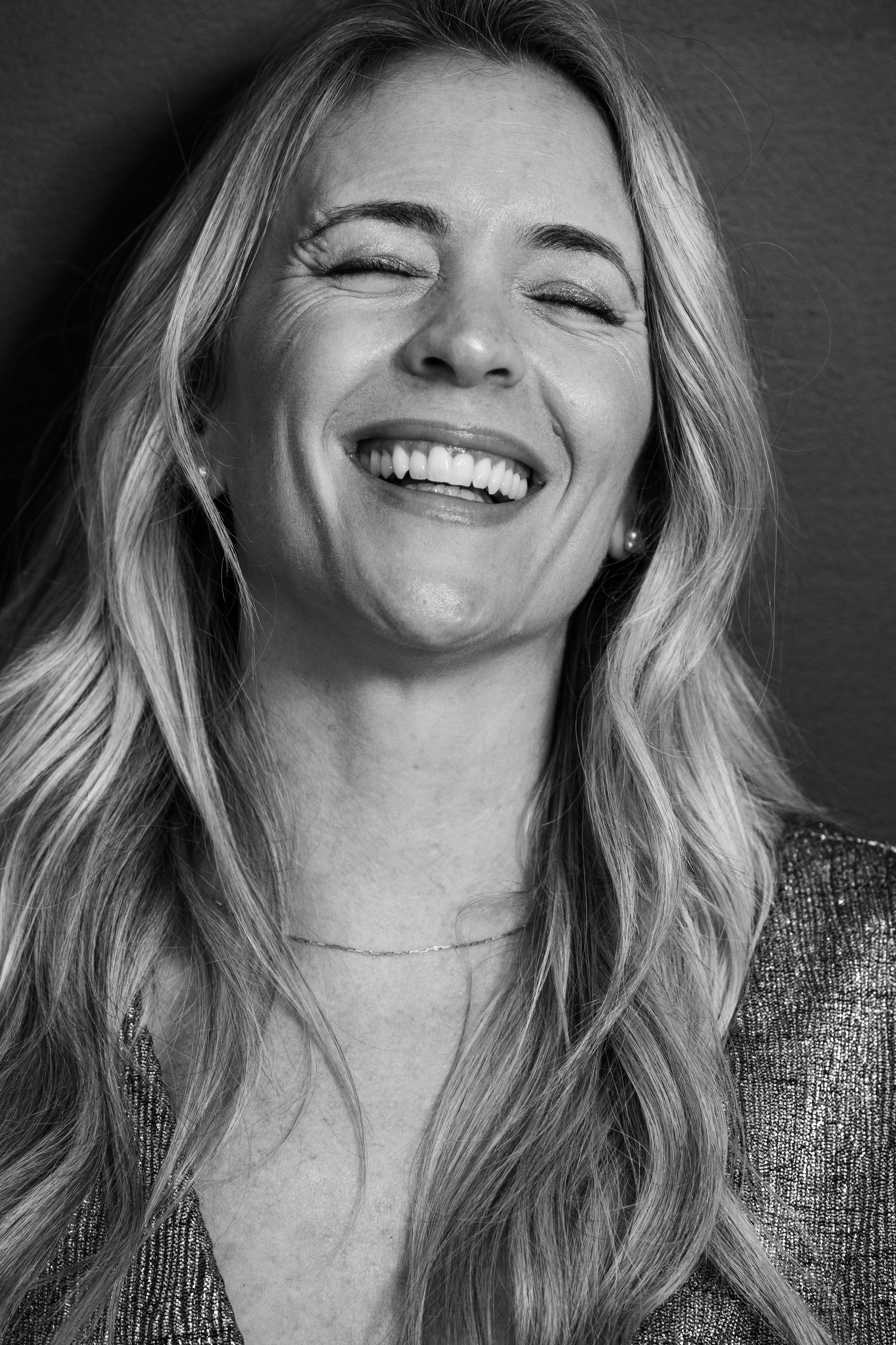 Black and white close-up portrait of a smiling woman with long wavy hair, wearing a textured top, earrings, and a necklace.
