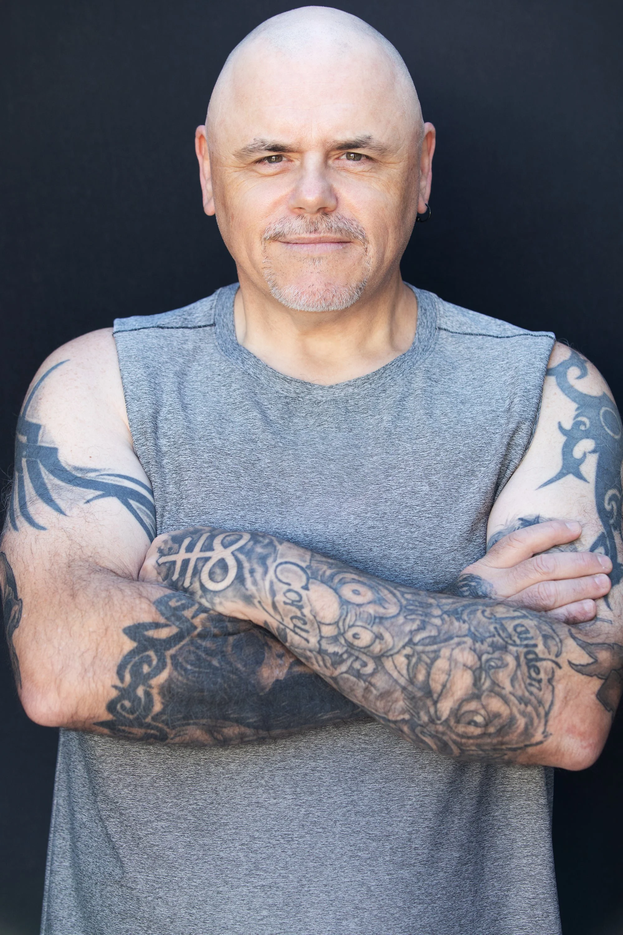 A bald man with a goatee, wearing a sleeveless gray shirt, crossing his arms and looking at the camera against a dark background.