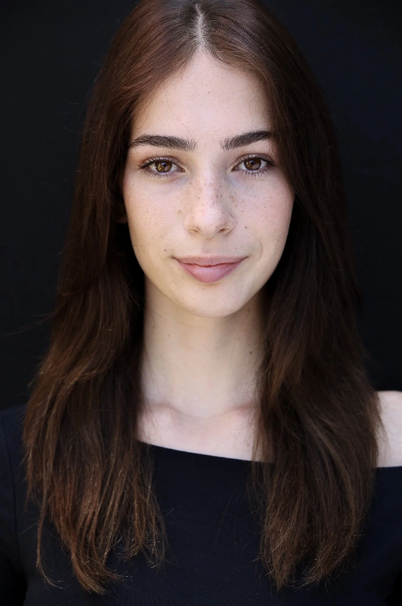 A young woman with long, wavy brown hair, light skin, freckles, and brown eyes, wearing a black top, smiling softly against a dark background.