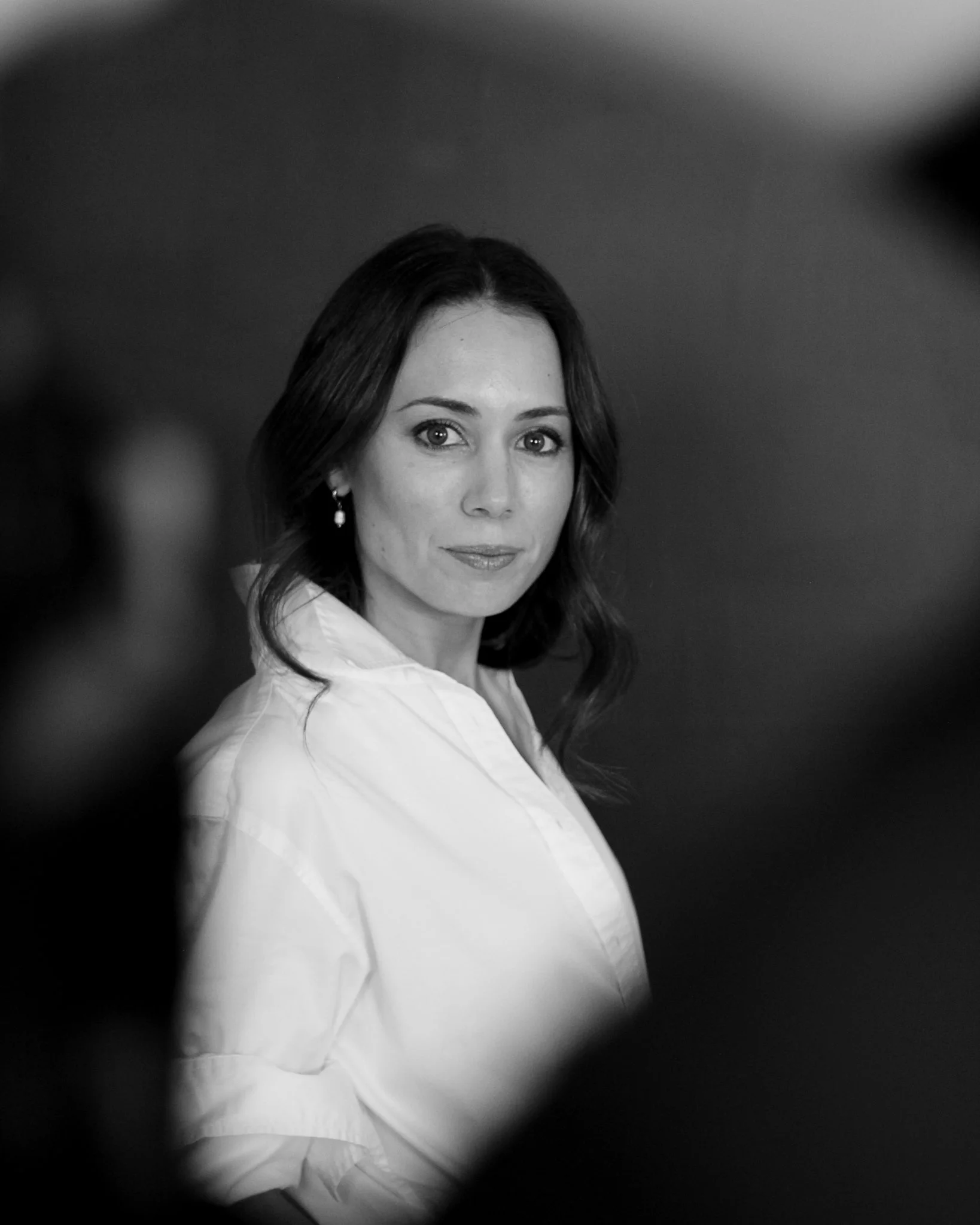Black and white headshot of a woman with shoulder-length hair, wearing earrings and a white shirt, looking at the camera with a neutral expression.