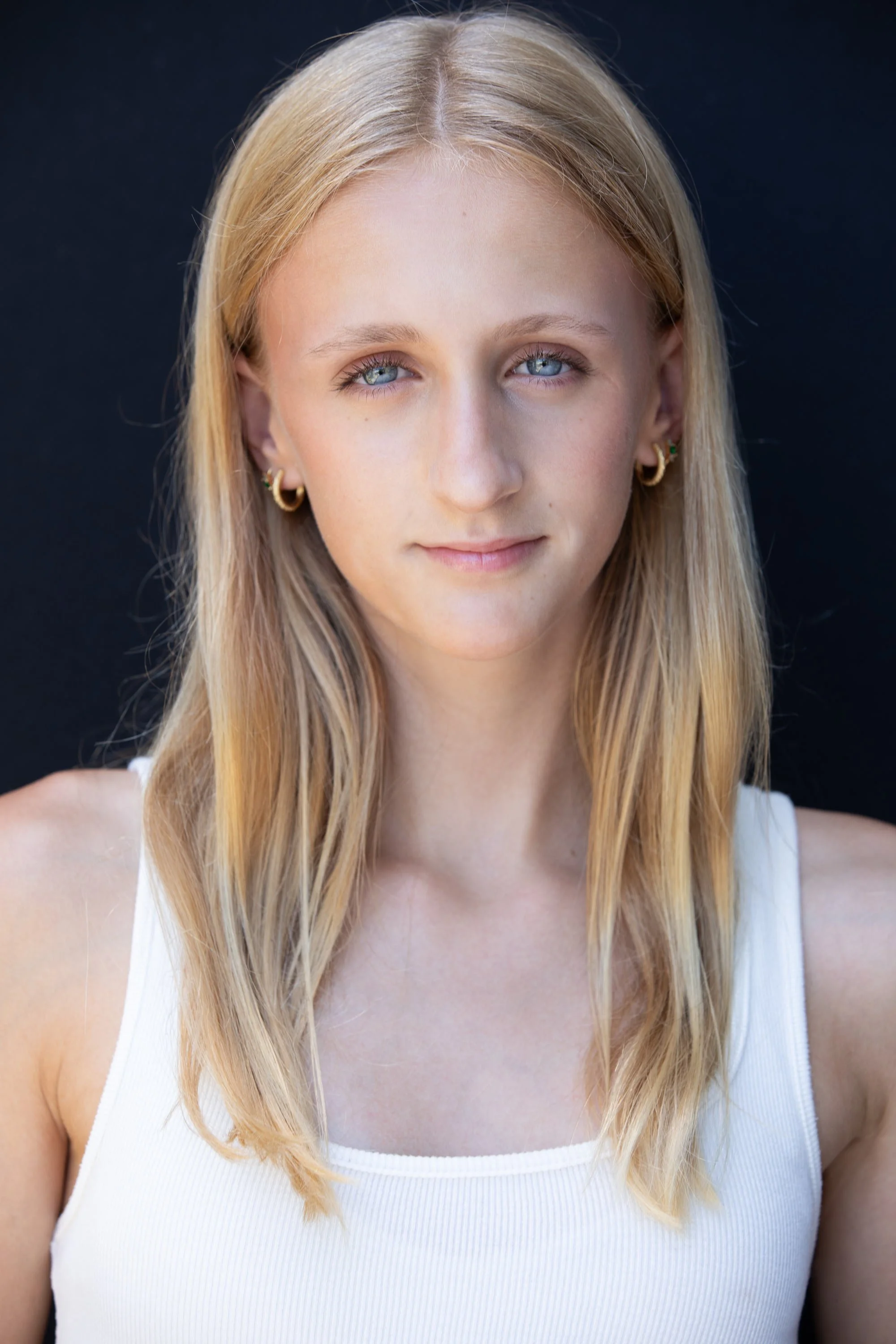 Close-up of a young woman with long blonde hair, blue eyes, gold hoop earrings, looking directly at the camera with a slight smile, wearing a white sleeveless top, against a dark background.