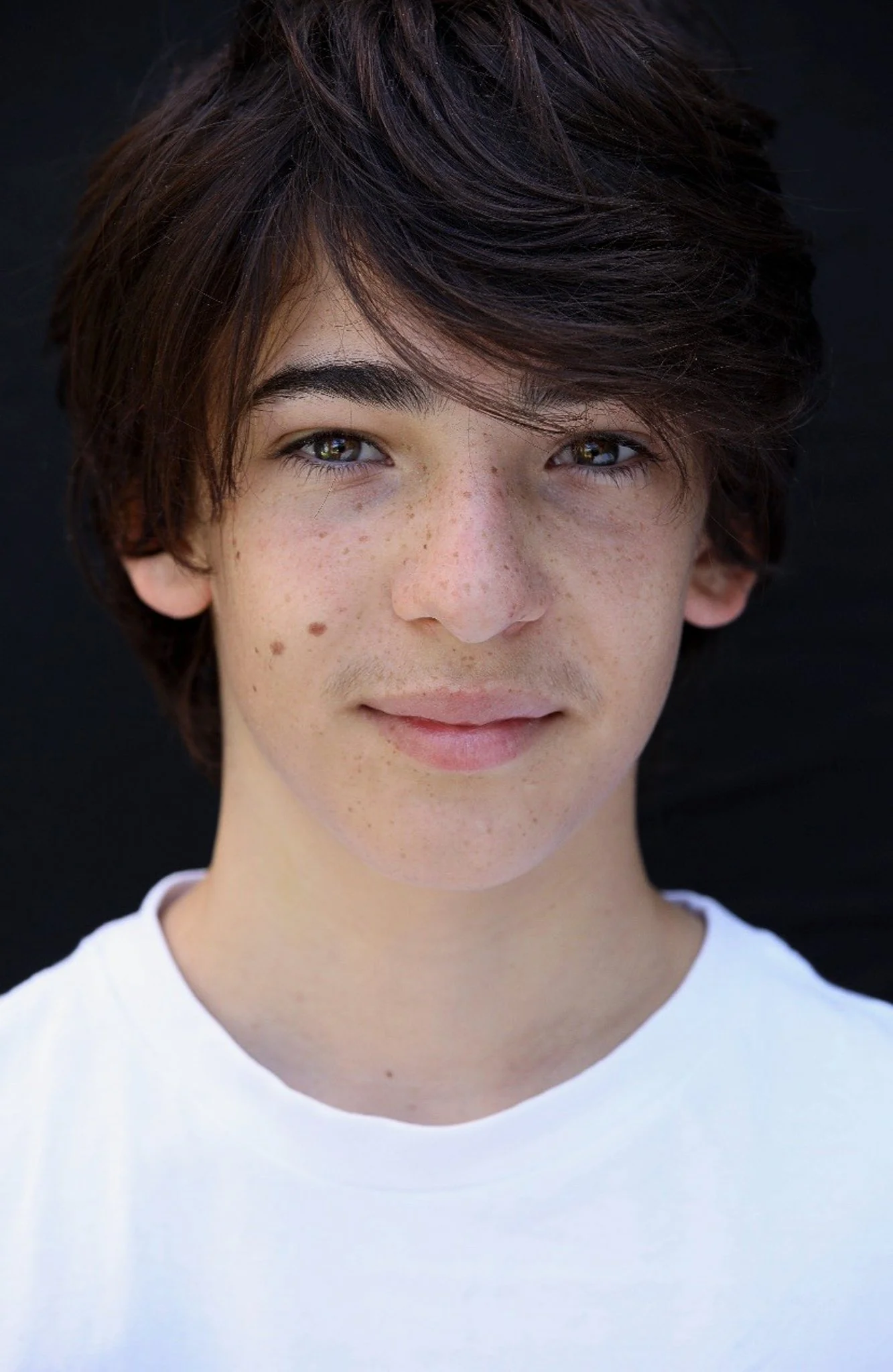 Close-up of a young person with dark brown hair, freckles, and hazel eyes, wearing a white shirt, against a black background.