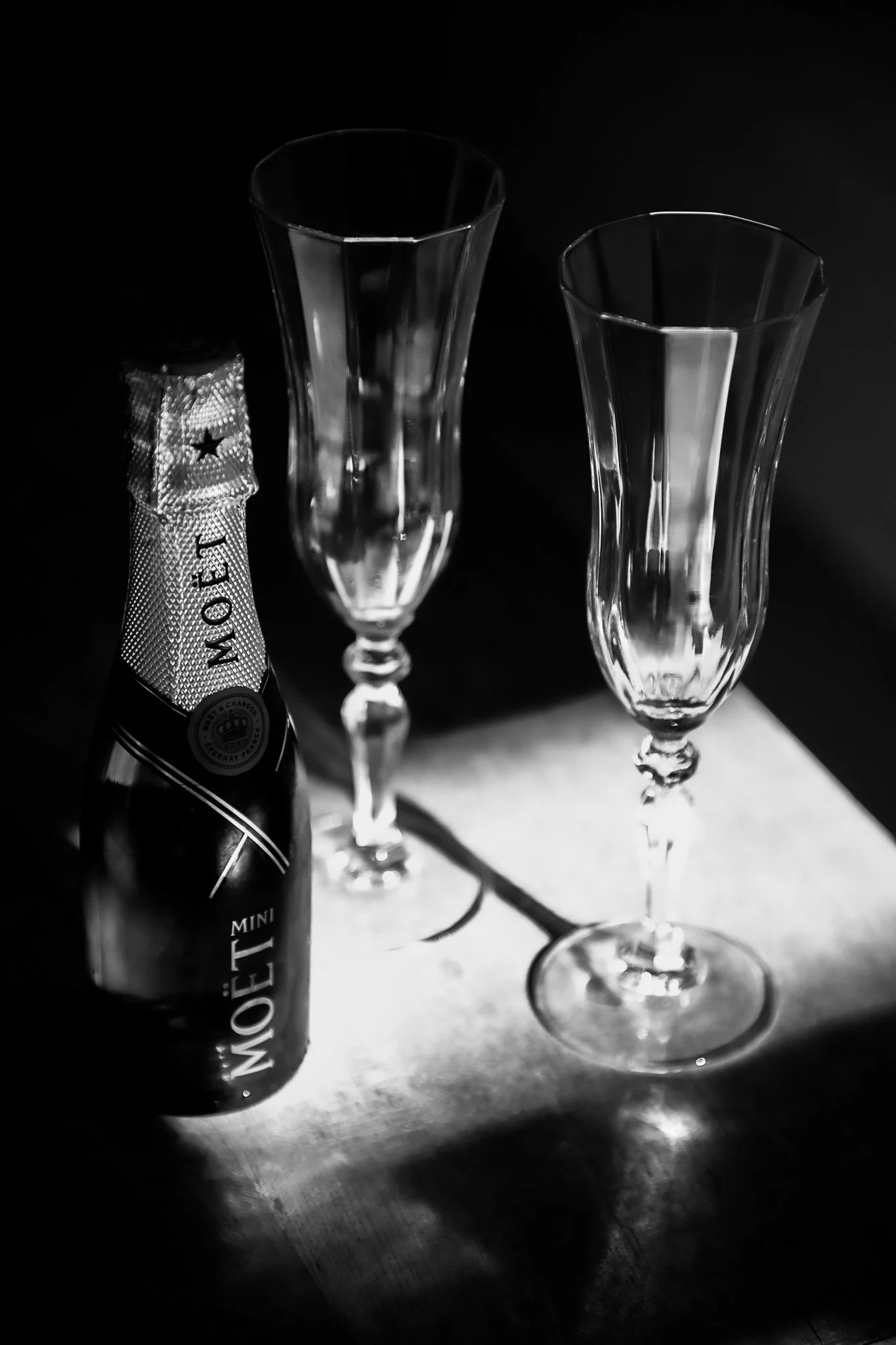 A bottle of Moët & Chandon champagne and two empty champagne flutes on a table in a black and white photo.