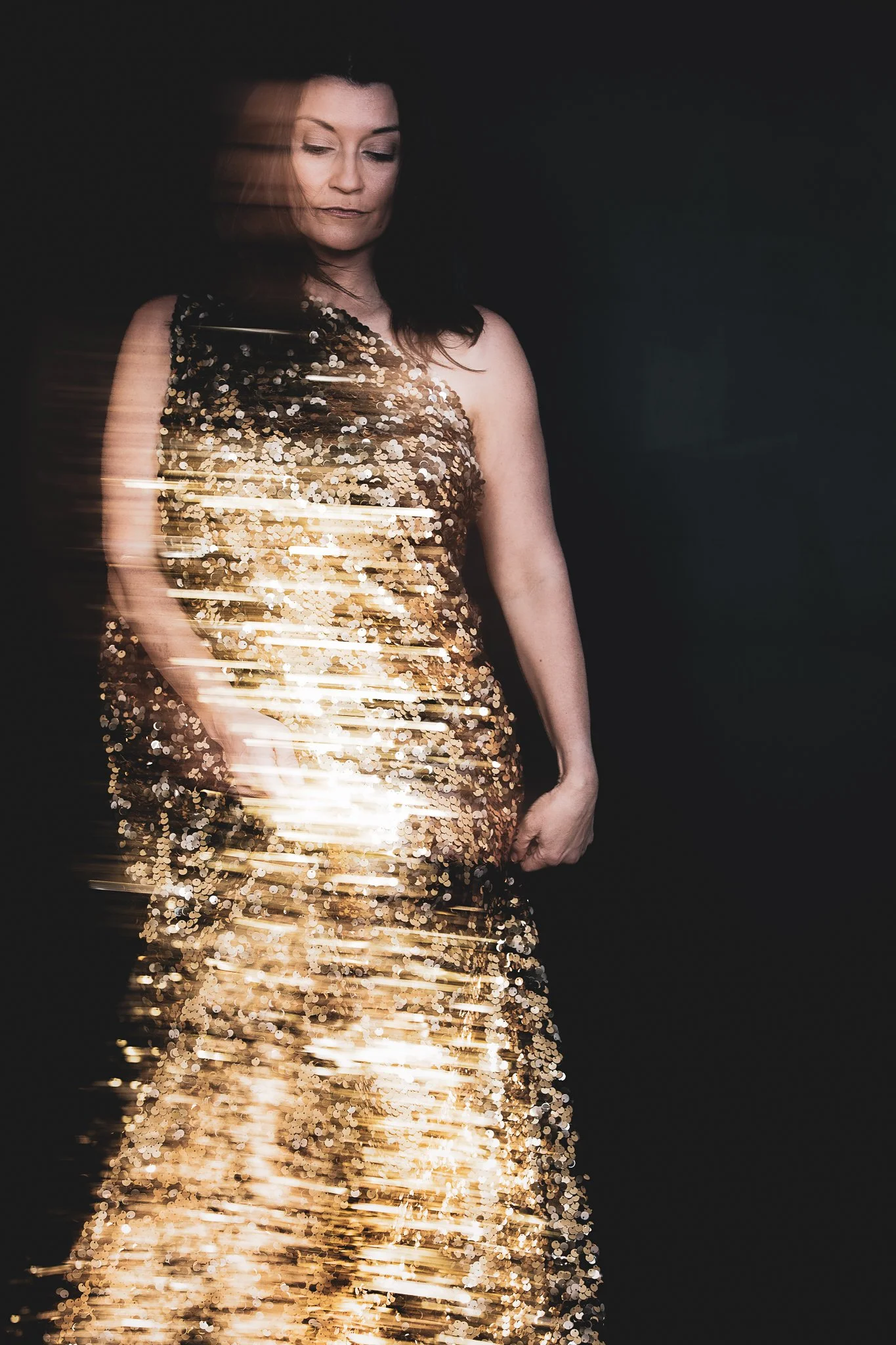 Woman in a gold sequin evening gown standing against a dark background, with motion blur effects.