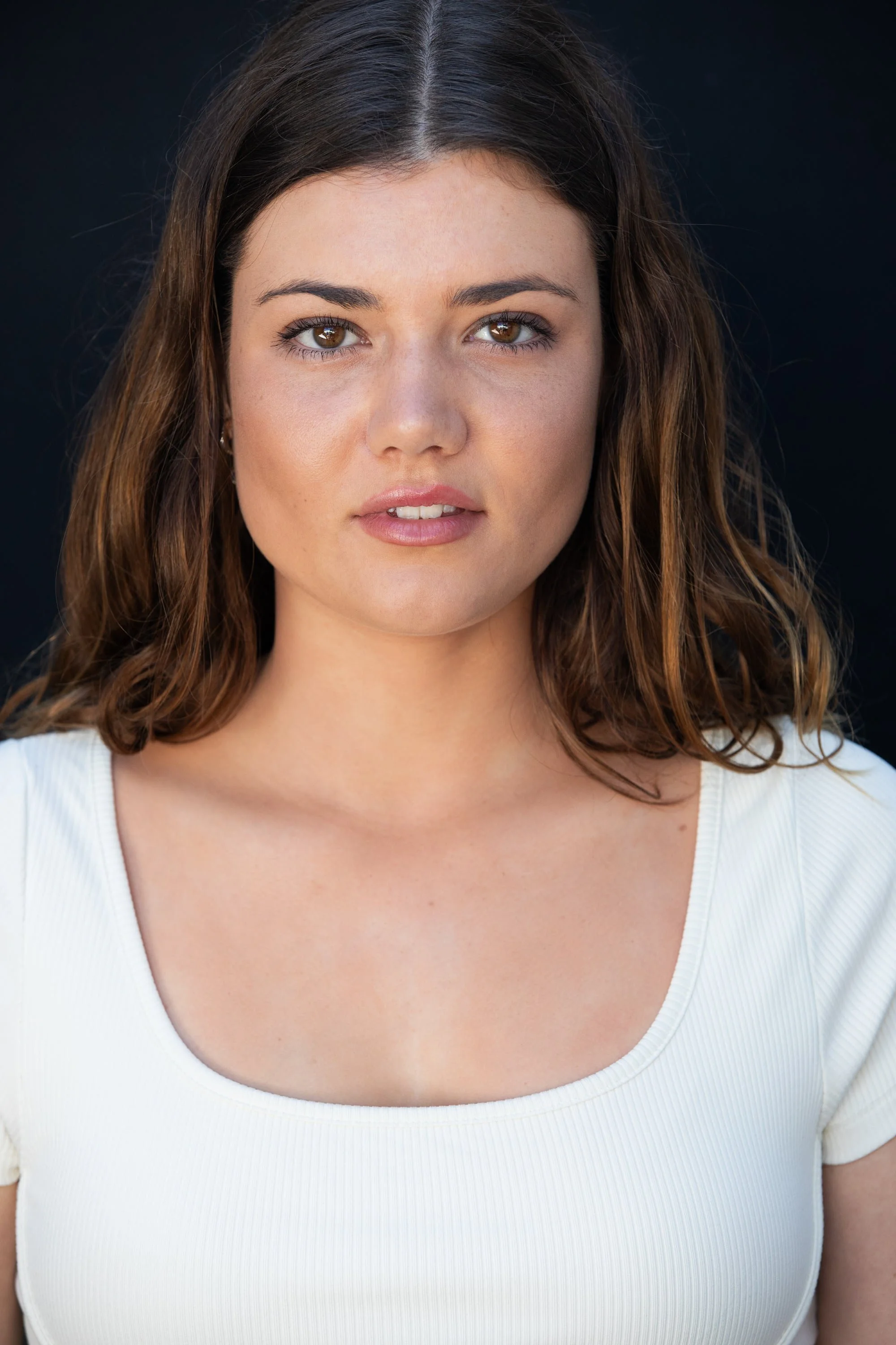 A woman with shoulder-length brown hair, brown eyes, and light skin wearing a white scoop-neck top, standing against a dark background.