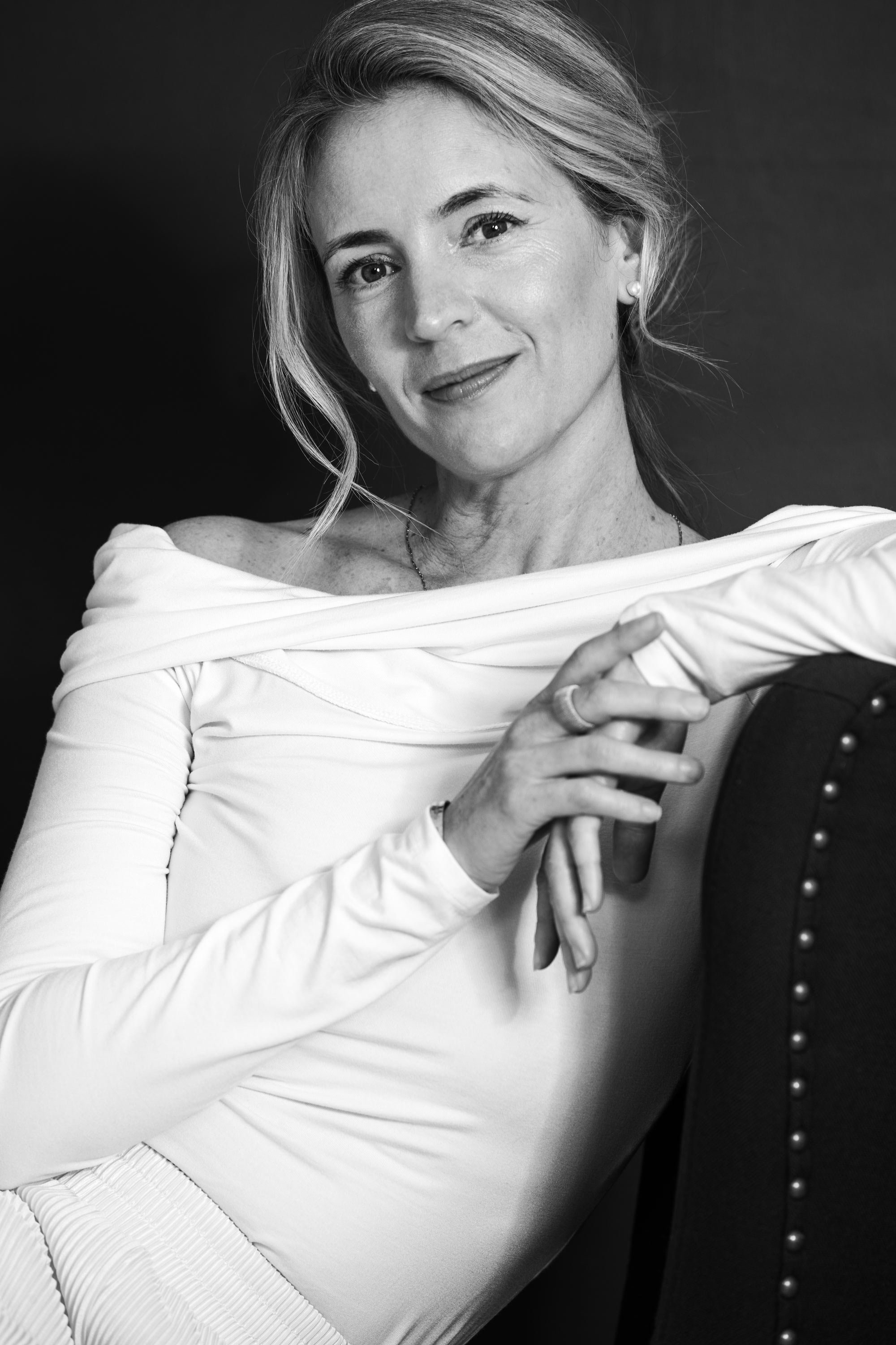 A black and white portrait of a woman smiling, with her left hand resting on her shoulder, wearing a white off-the-shoulder top and pearl earrings.