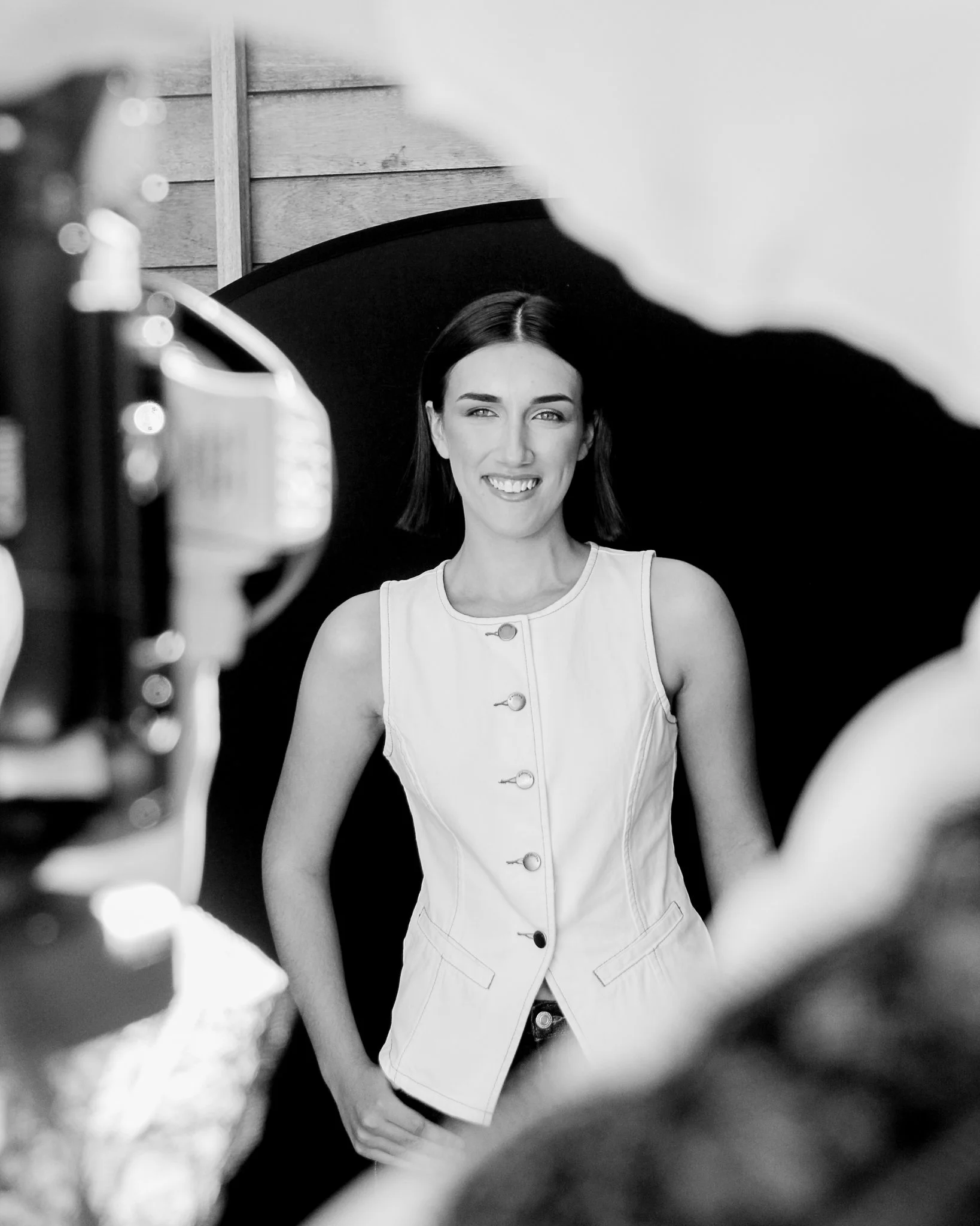 Black and white photo of a woman smiling, visible through a circular object in the foreground, with a wooden wall background.