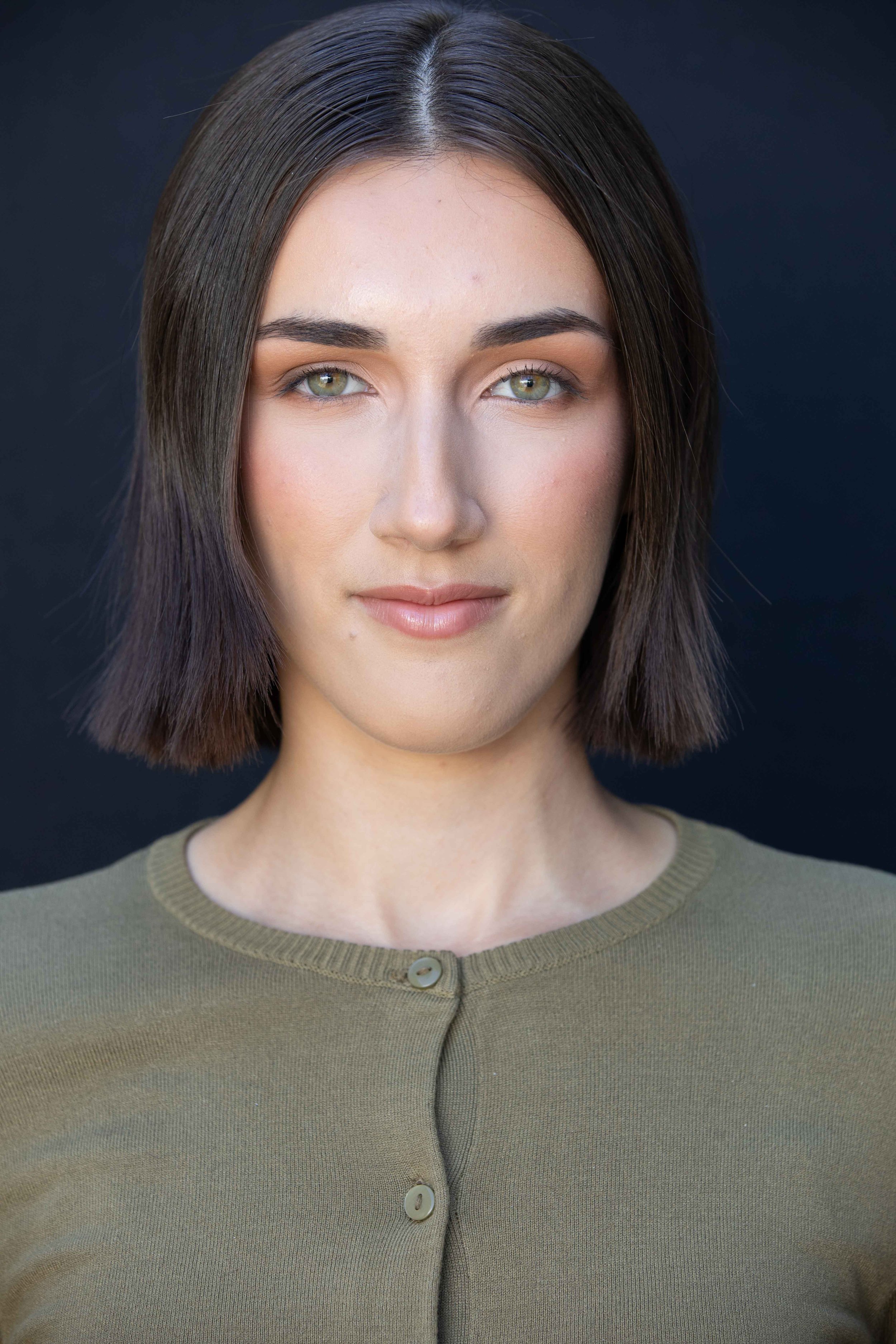 Close-up portrait of a woman with shoulder-length dark brown hair, green eyes, and a neutral expression, wearing a buttoned olive green top against a solid dark background.