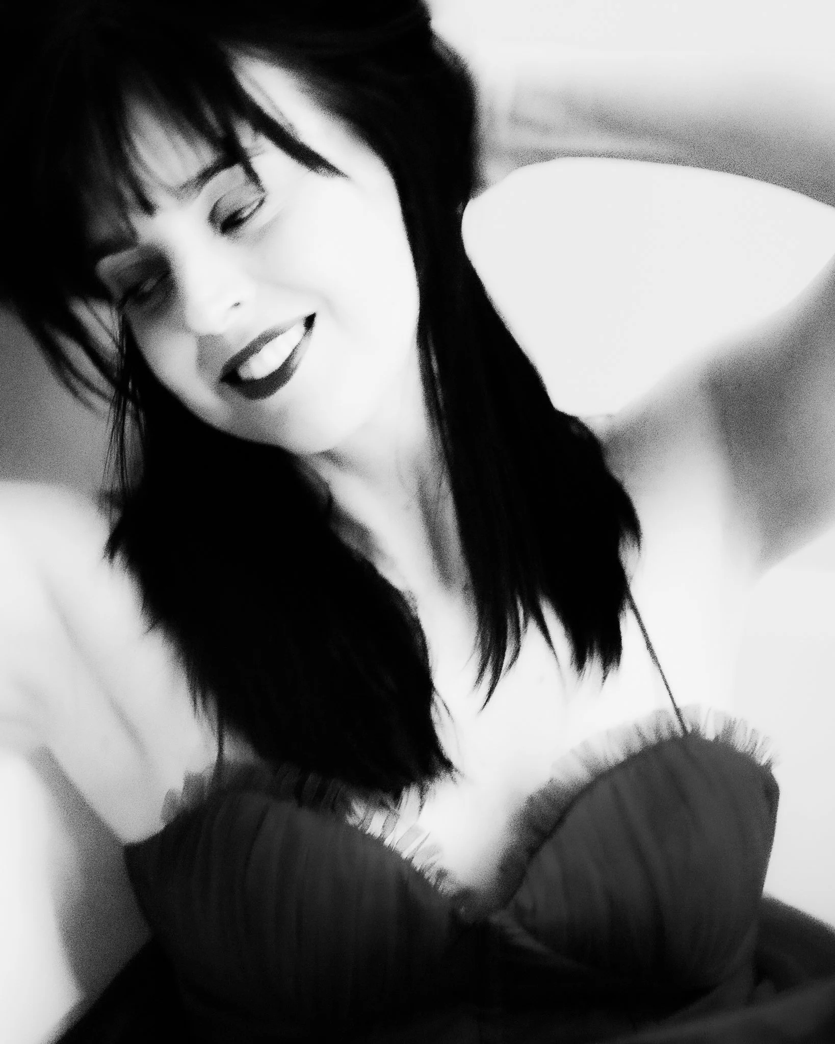 Black and white photo of a woman smiling, with her eyes closed, hair covering part of her face, and holding her arm near her face.