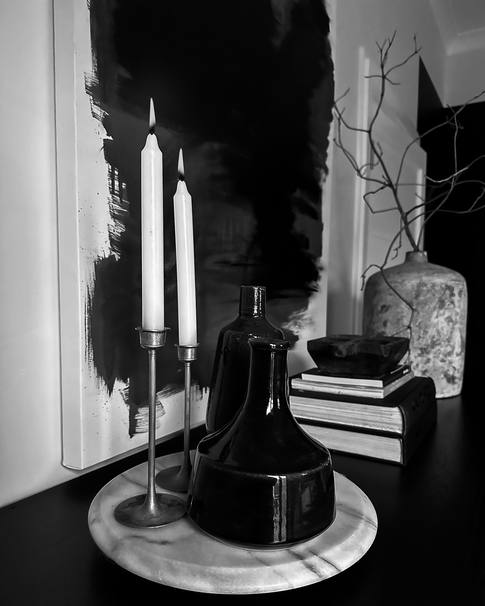 Black and white photo of a shelf with two lit candles, a dark glass bottle, a stack of books, and a decorative urn. A framed abstract painting with black brushstrokes is behind the objects, and a branch with leaves in a vase is on the right side.