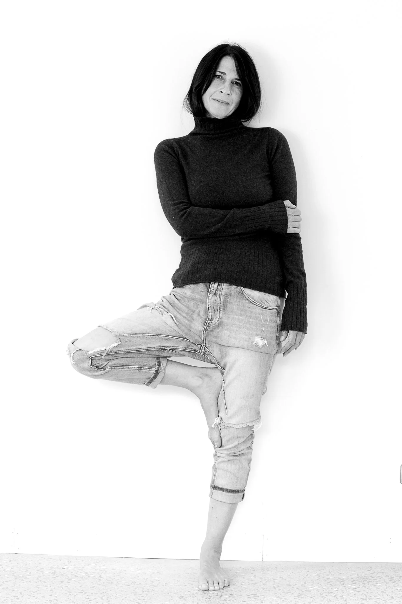 Black and white photo of a woman standing against a white wall, with her arms crossed, wearing a black turtleneck sweater and torn jeans, one leg lifted with foot resting against the wall.