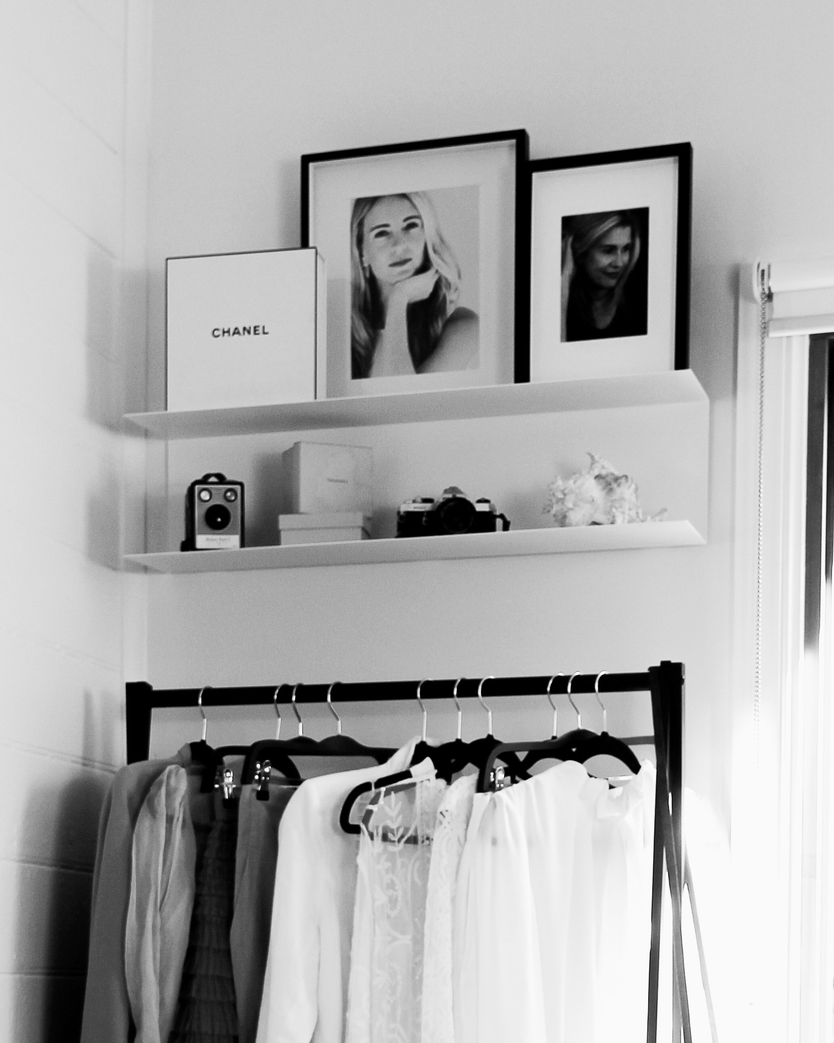 Black and white photo of a room with a shelf holding framed portraits, a book with 'Chanel' on the cover, a speaker, and a camera; below, a clothes rack with hanging garments.