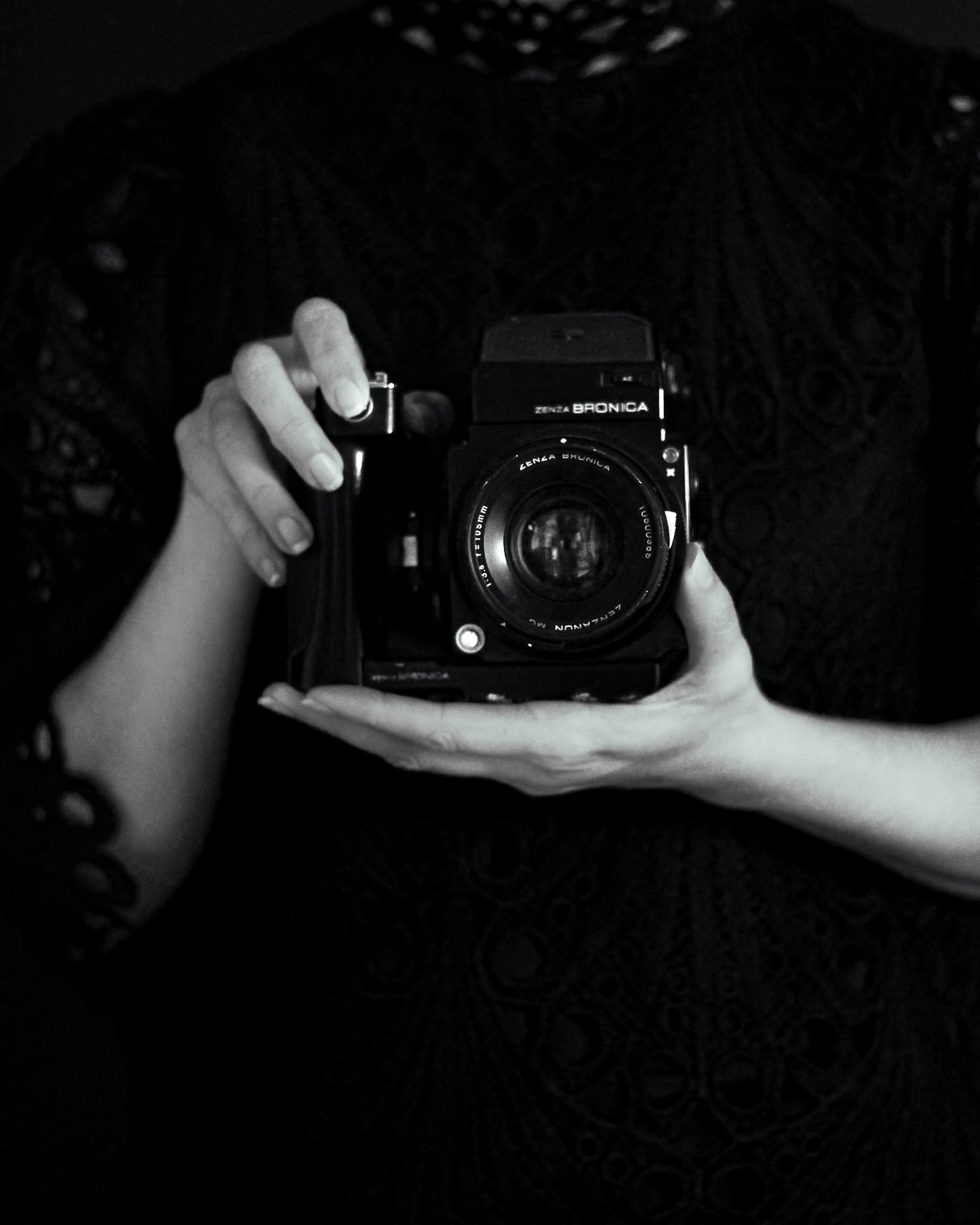 A person taking a selfie with a camera in a dark setting, only their hands and the camera are visible, with the camera brand 'Zenza Bronica' visible on the camera body.