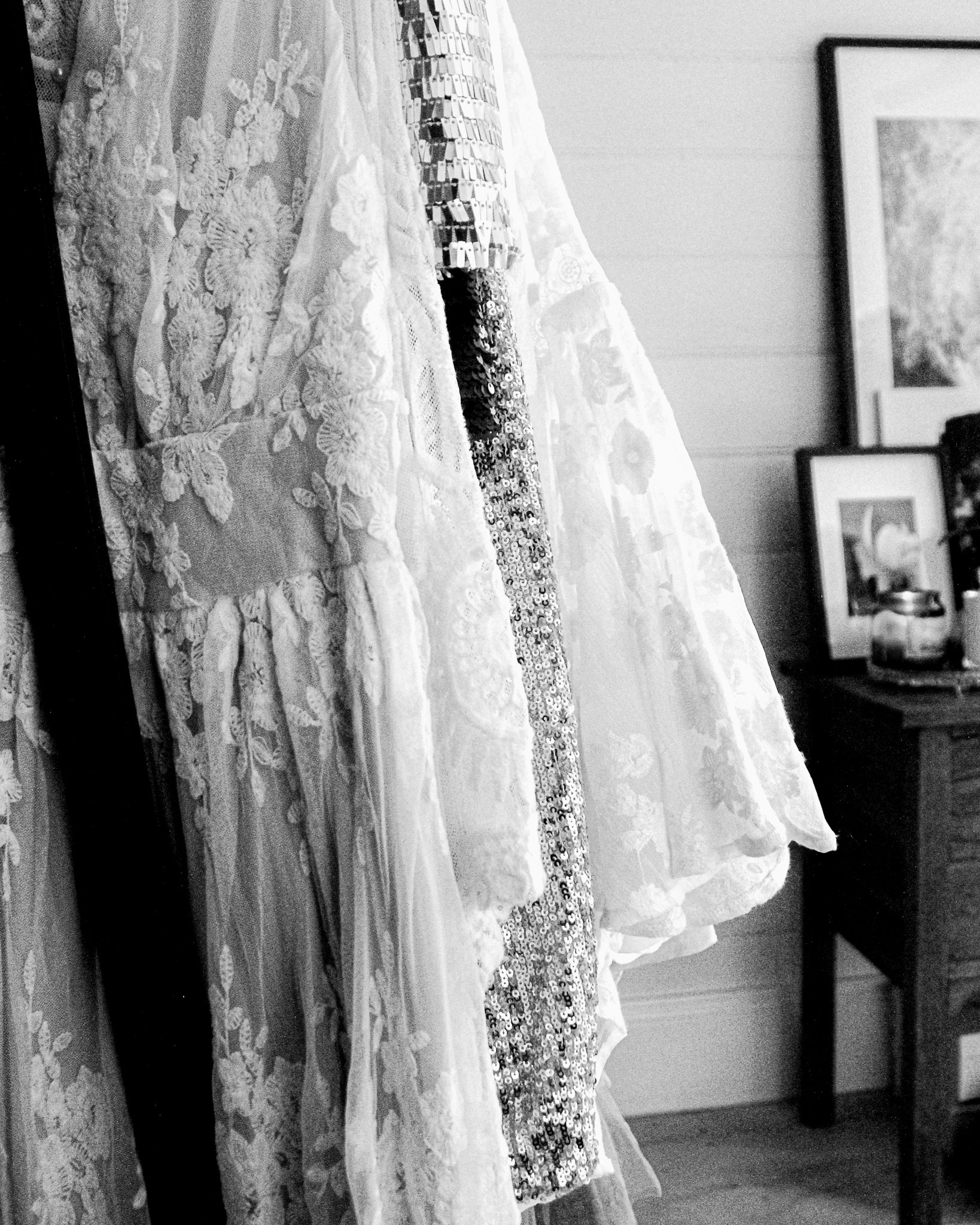 Close-up of a lace dress with intricate embroidery and beaded embellishments, hanging in a room with framed pictures on the wall in the background.