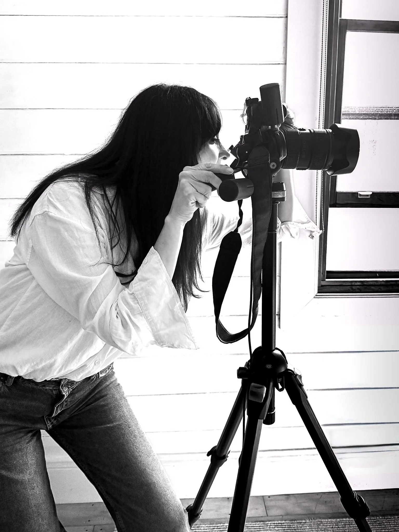 A woman with long dark hair wearing a white shirt and jeans is kneeling, looking through the viewfinder of a camera mounted on a tripod.