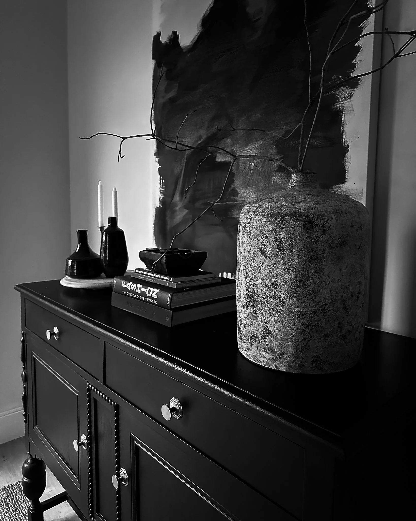 A black wooden sideboard with five drawers, brass knobs, and turned legs, topped with three black bottles, a white round tray, a stack of books, and a large decorative vase, with a large canvas painting of a horse in neutral tones hanging above.