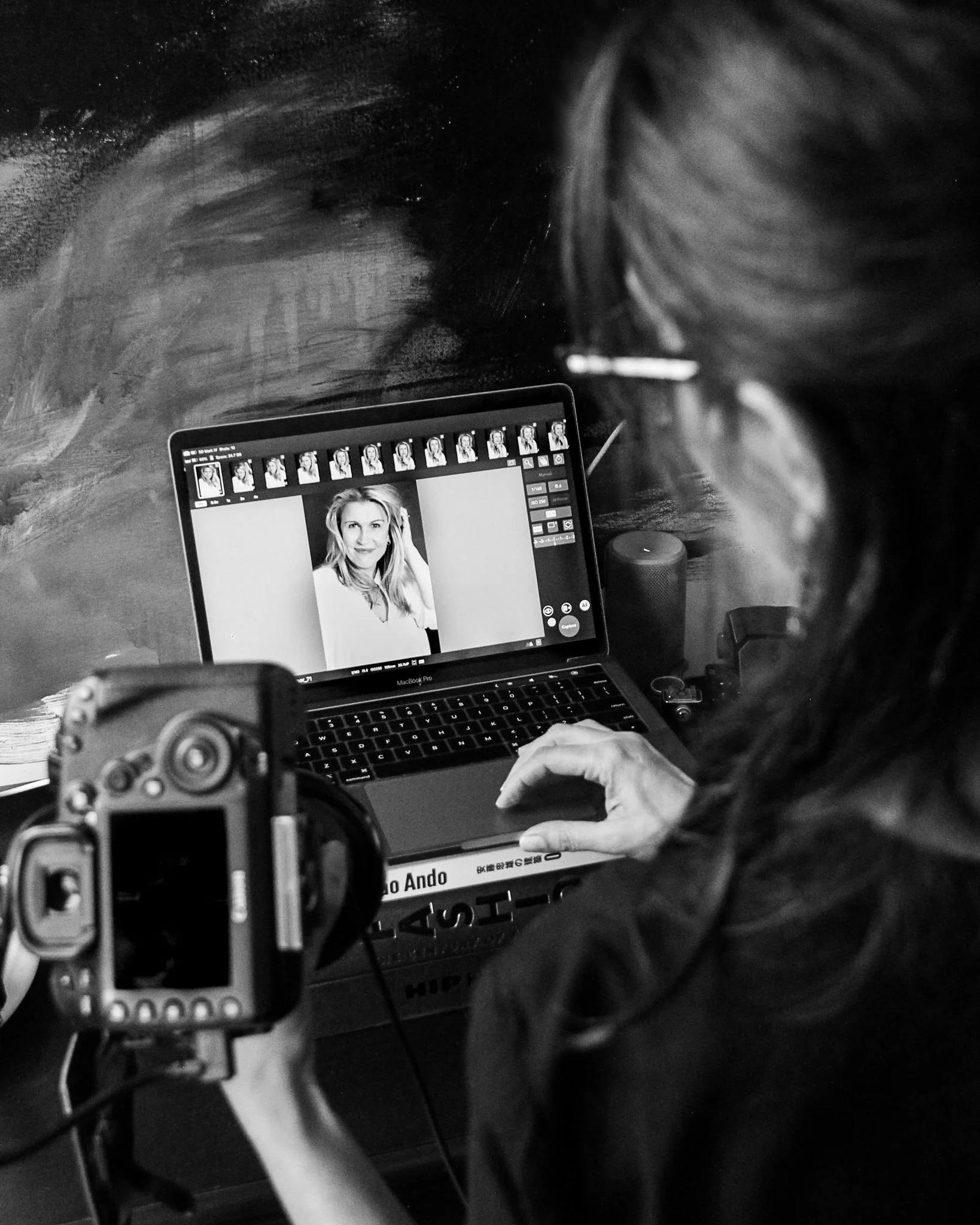 A person standing at a desk with a laptop displaying a black-and-white portrait photo of a woman with blonde hair. The person is also holding a camera. The background features a textured wall.