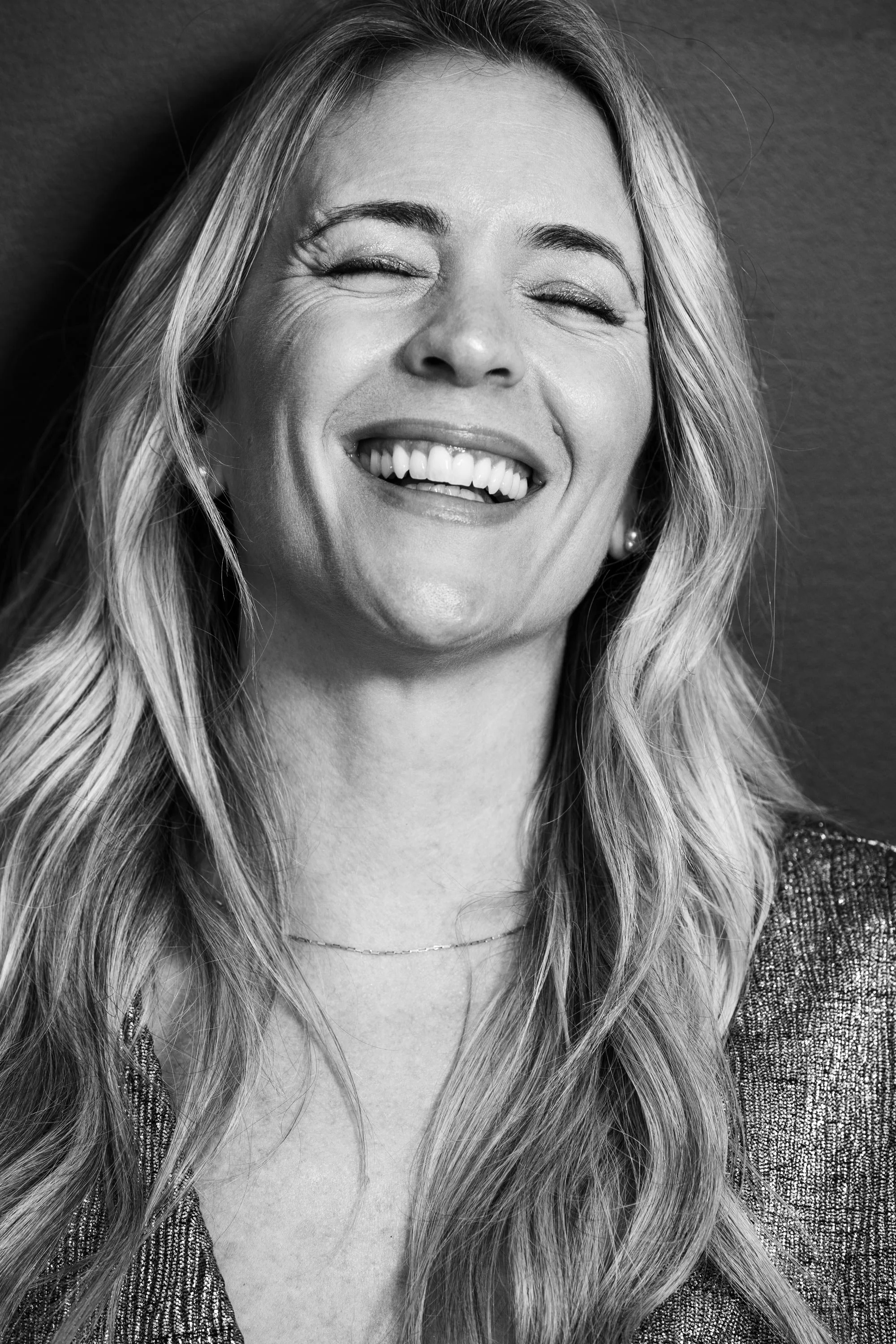 Black and white close-up of a smiling woman with long wavy hair, wearing a sparkly jacket, pearl earring, and a thin necklace.