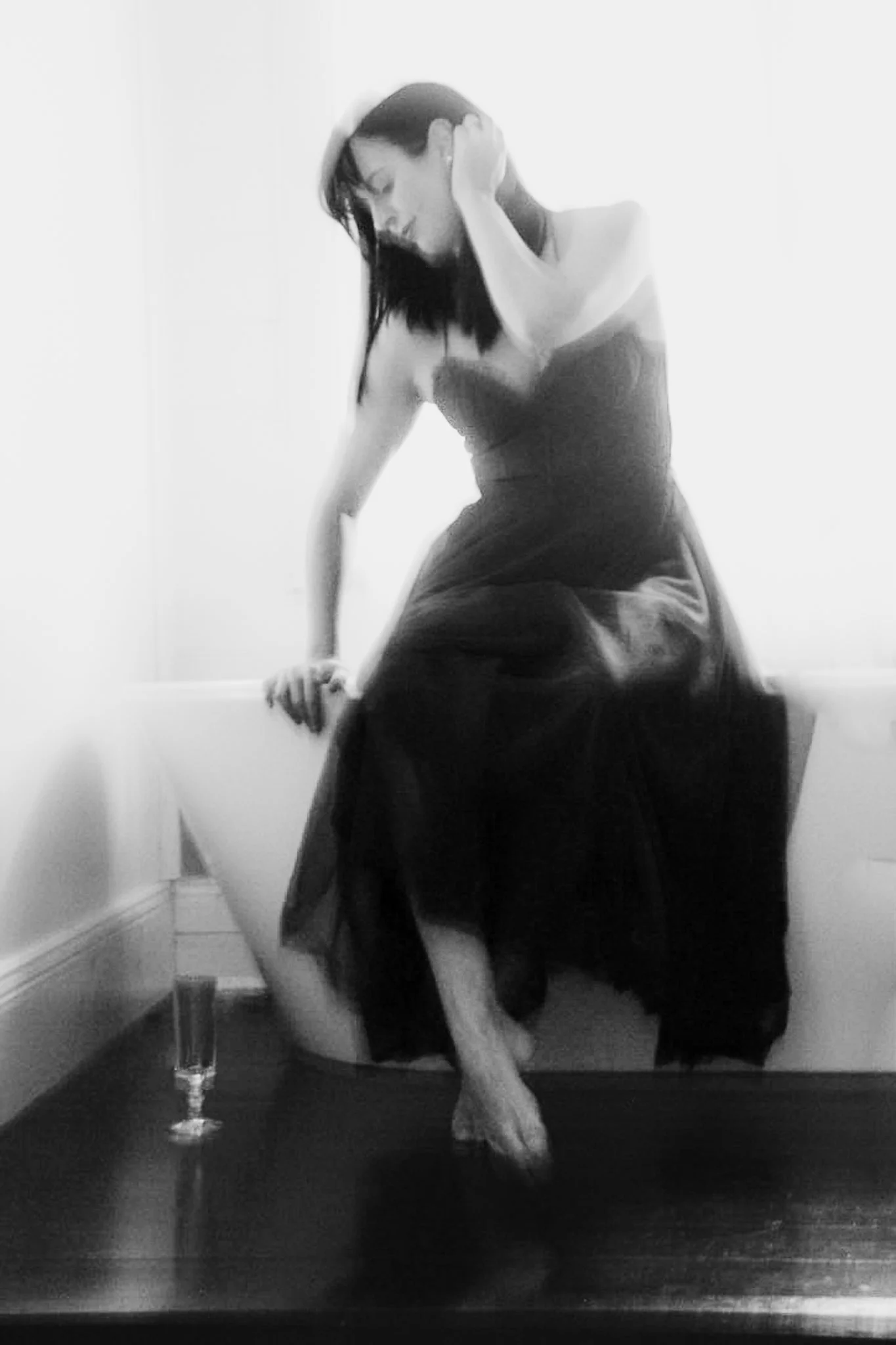 A woman with dark hair in a black strapless dress sitting on a chair in a room, with a glass of drink on a table next to her, positioned in front of a mirror.