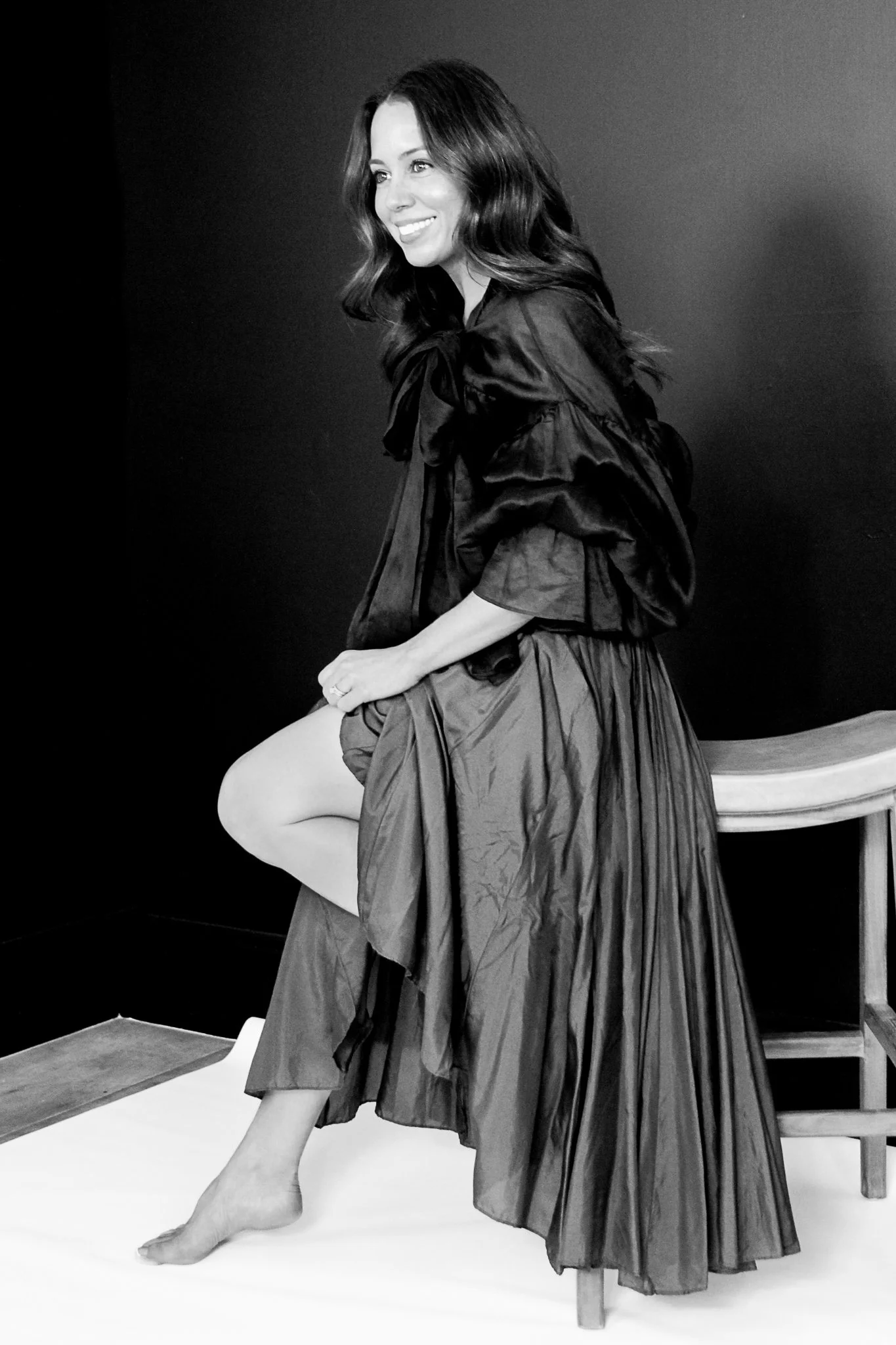 A woman with long wavy hair, smiling, seated on a wooden bench, wearing a dark, flowy dress and her leg is showing, in front of a dark background.