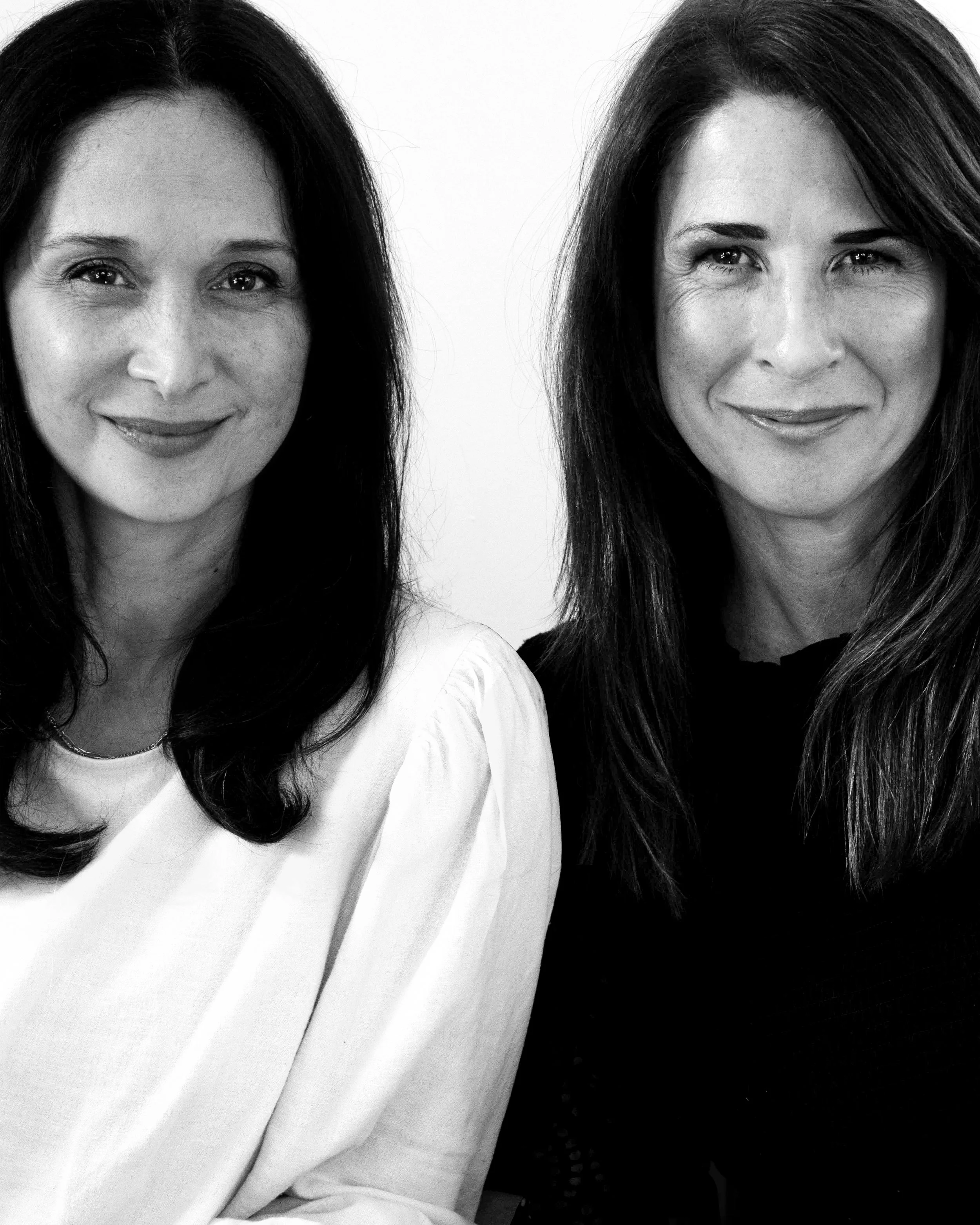 Black and white photo of two women smiling, one with dark hair and the other with slightly lighter hair, close-up headshots with plain background.