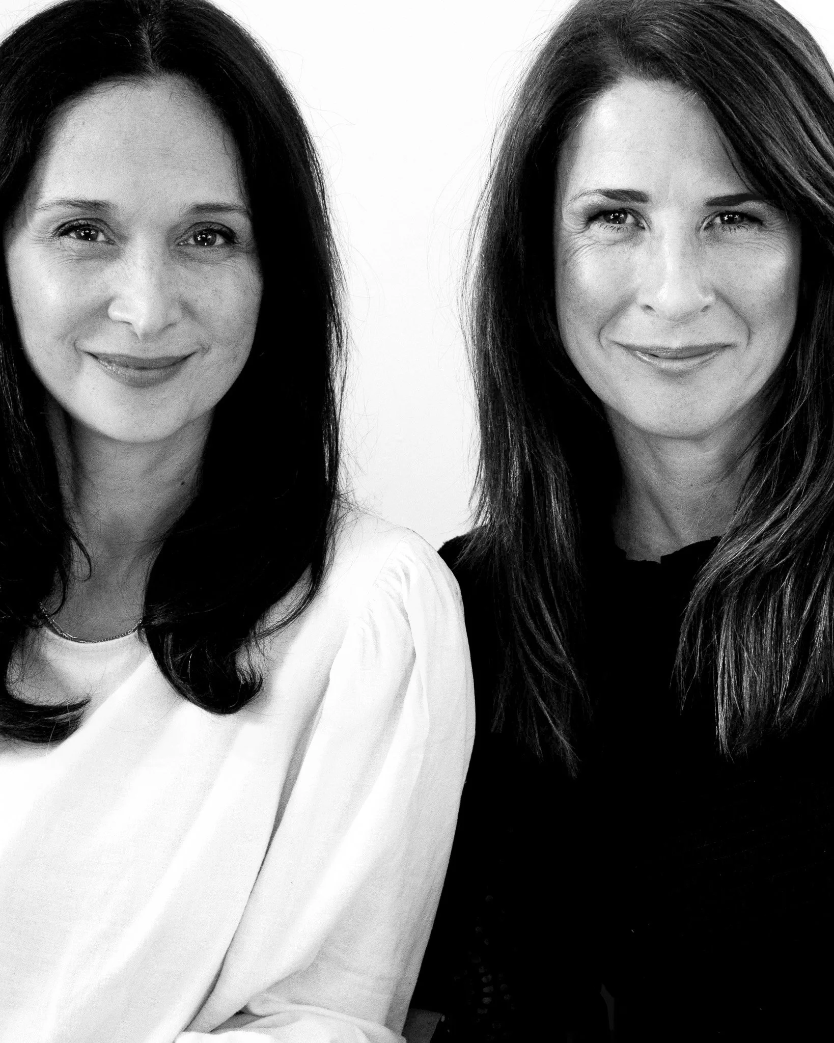 Black and white portrait of two women smiling. One woman has long dark hair and is wearing a light-colored top, the other has long dark hair and is wearing a dark top. The women are close together, and the photo captures their faces and upper bodies.