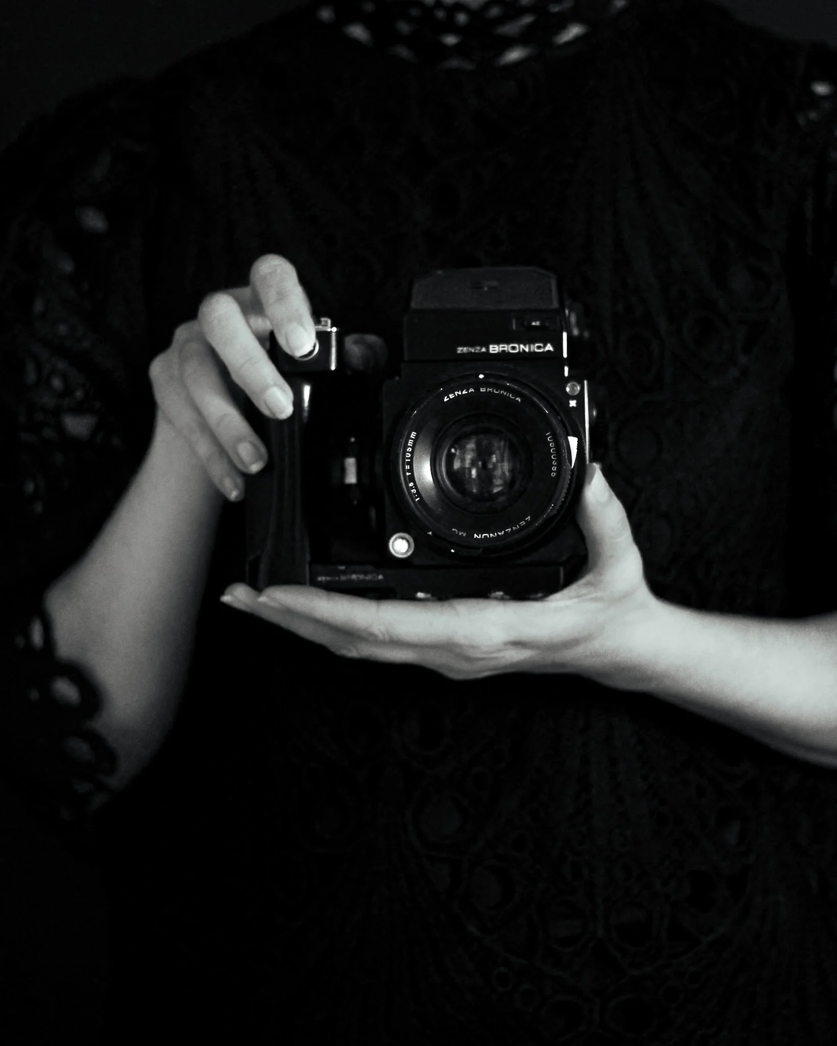 Person holding a vintage Zenza Bronica camera with the lens facing forward, partially visible hands, dark background.