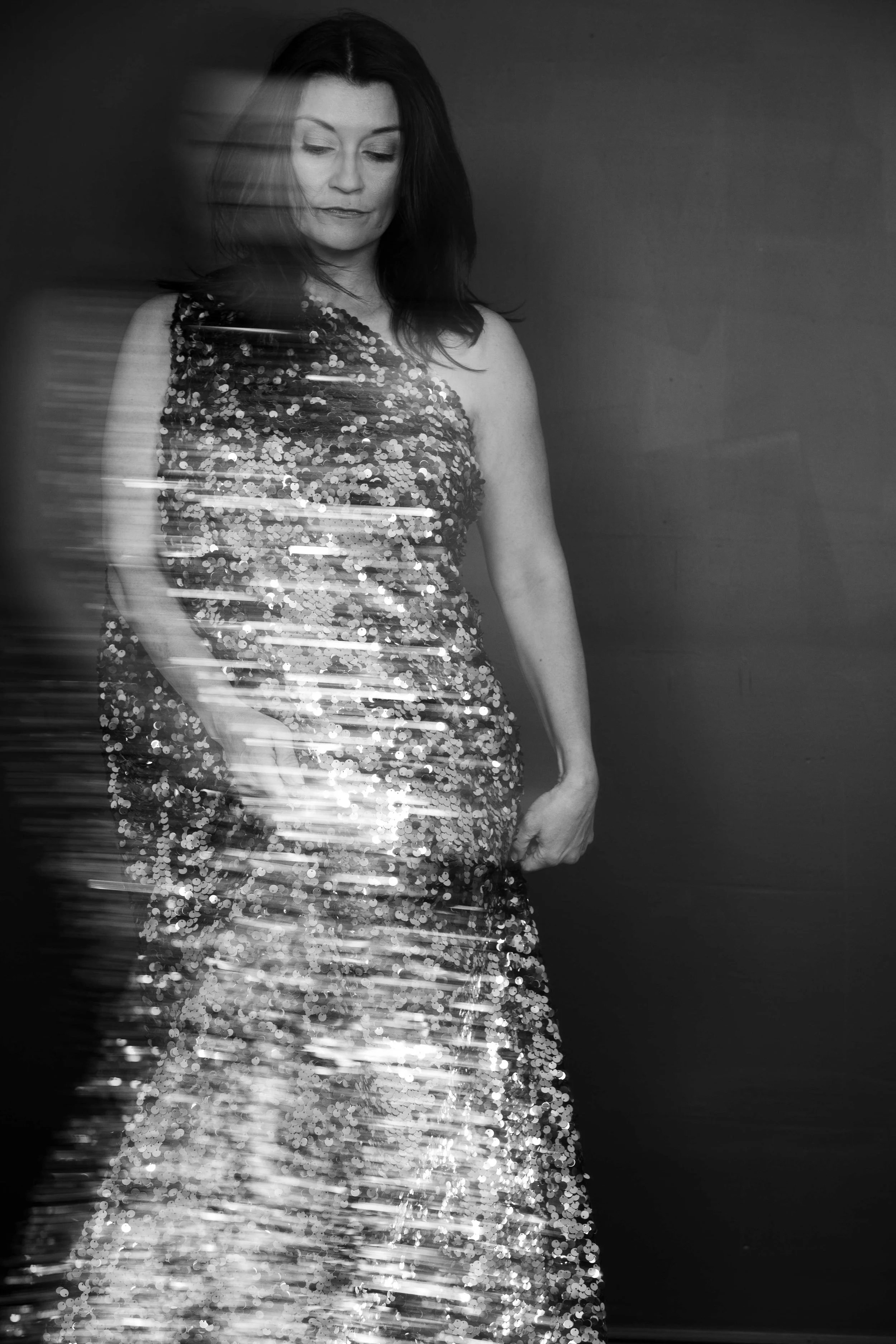 Black and white photo of a woman in a sleeveless, sequined dress, looking downward with a calm expression, her hair down, against a plain background.