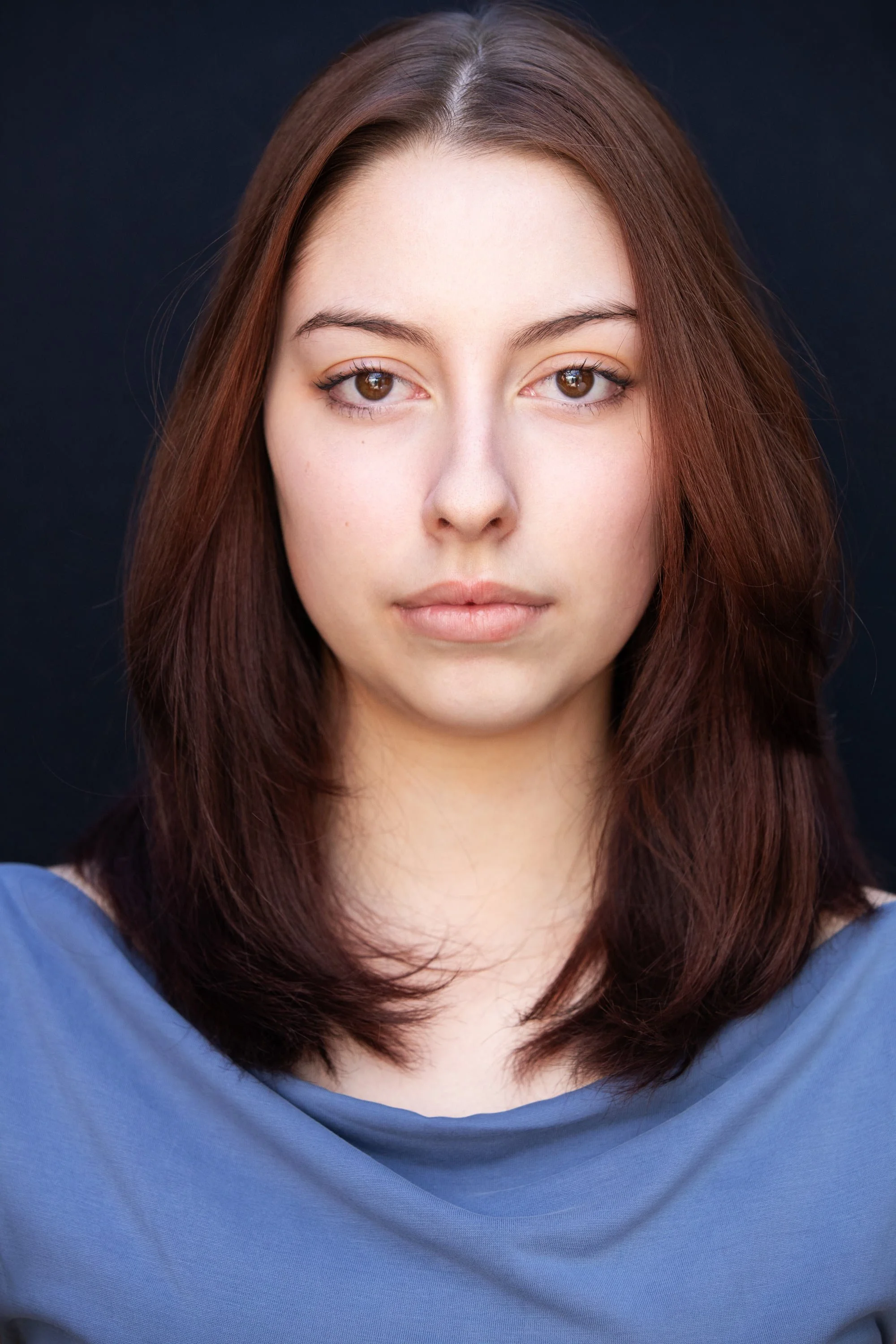 Close-up portrait of a woman with brown hair and brown eyes, wearing a blue top, against a dark background.