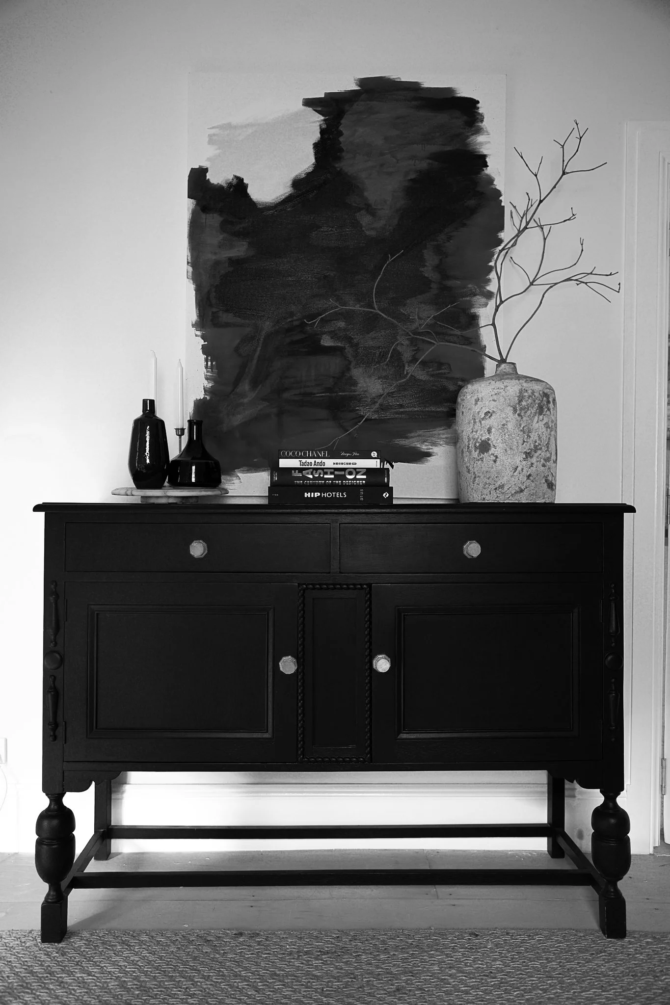 A black wooden sideboard with decorative molding, featuring a round marble tray with dark vases and candlesticks, with a stack of books on top. Behind the sideboard, a large abstract black and white painting hangs on the wall, with a tall ceramic vas