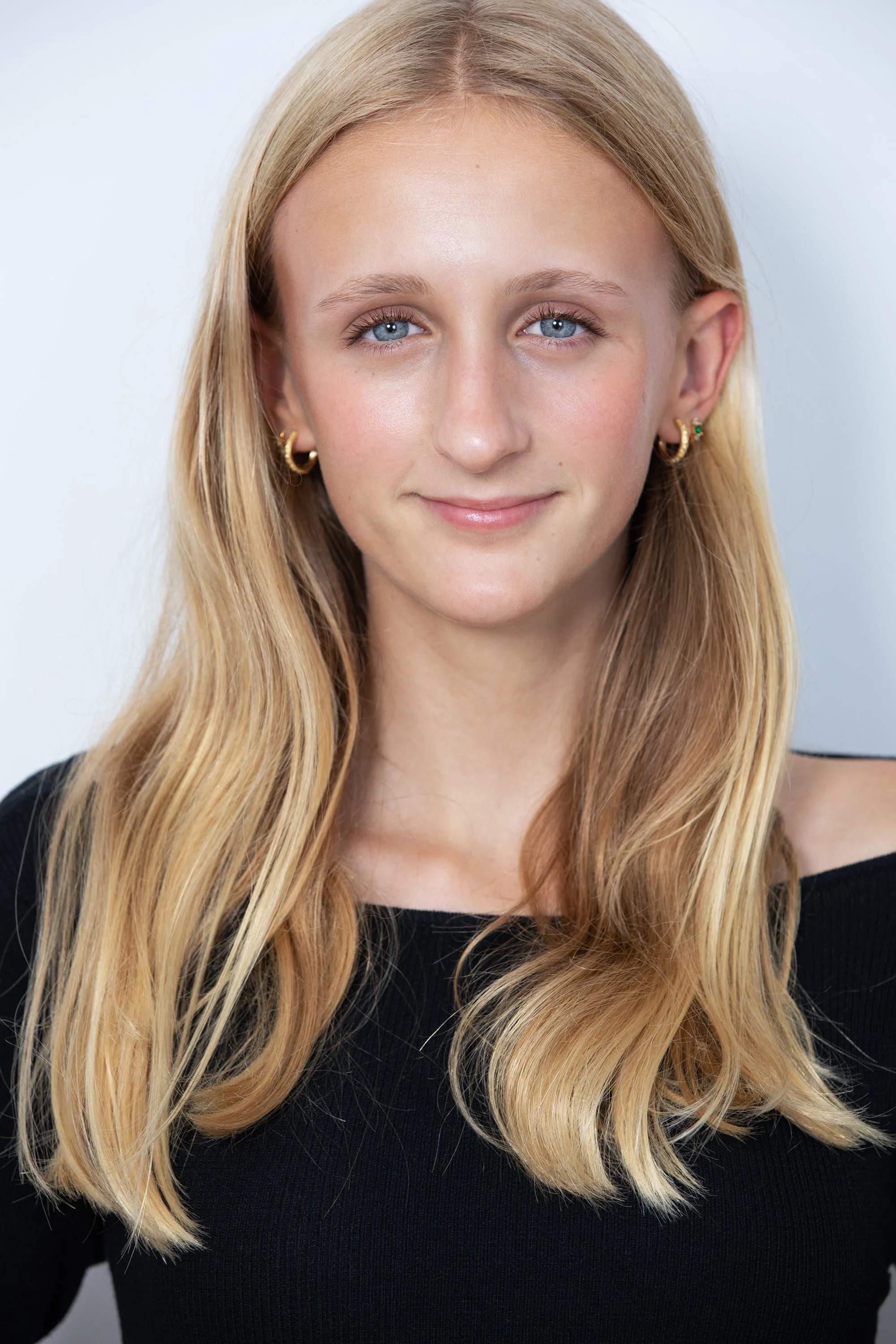 A young woman with long, wavy blonde hair, blue eyes, wearing gold hoop earrings and a black top, smiling softly against a plain light background.