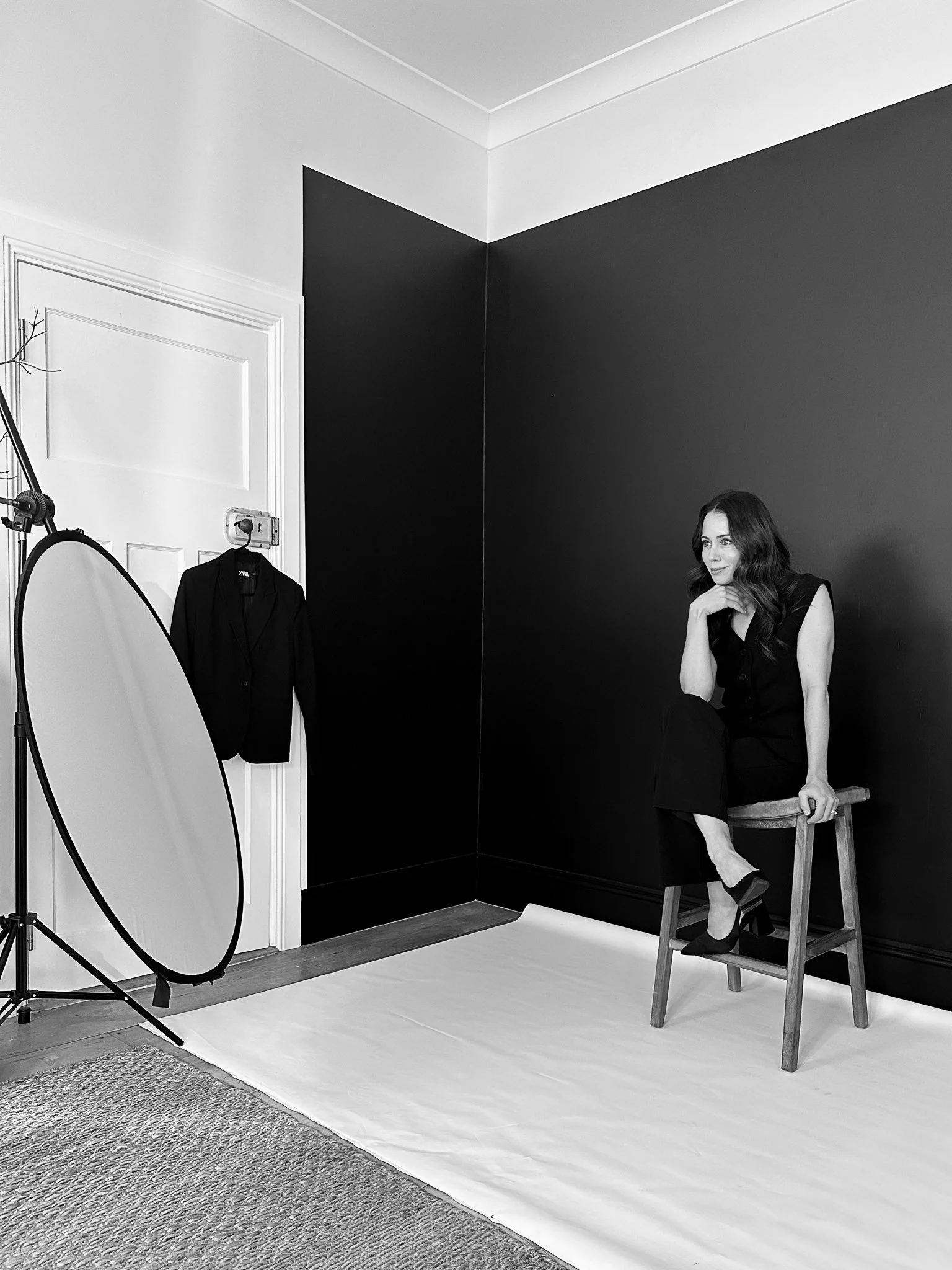 A woman in a sleeveless black dress sitting on a wooden chair, posing with one leg crossed over the other, inside a photography studio with a black wall background.
