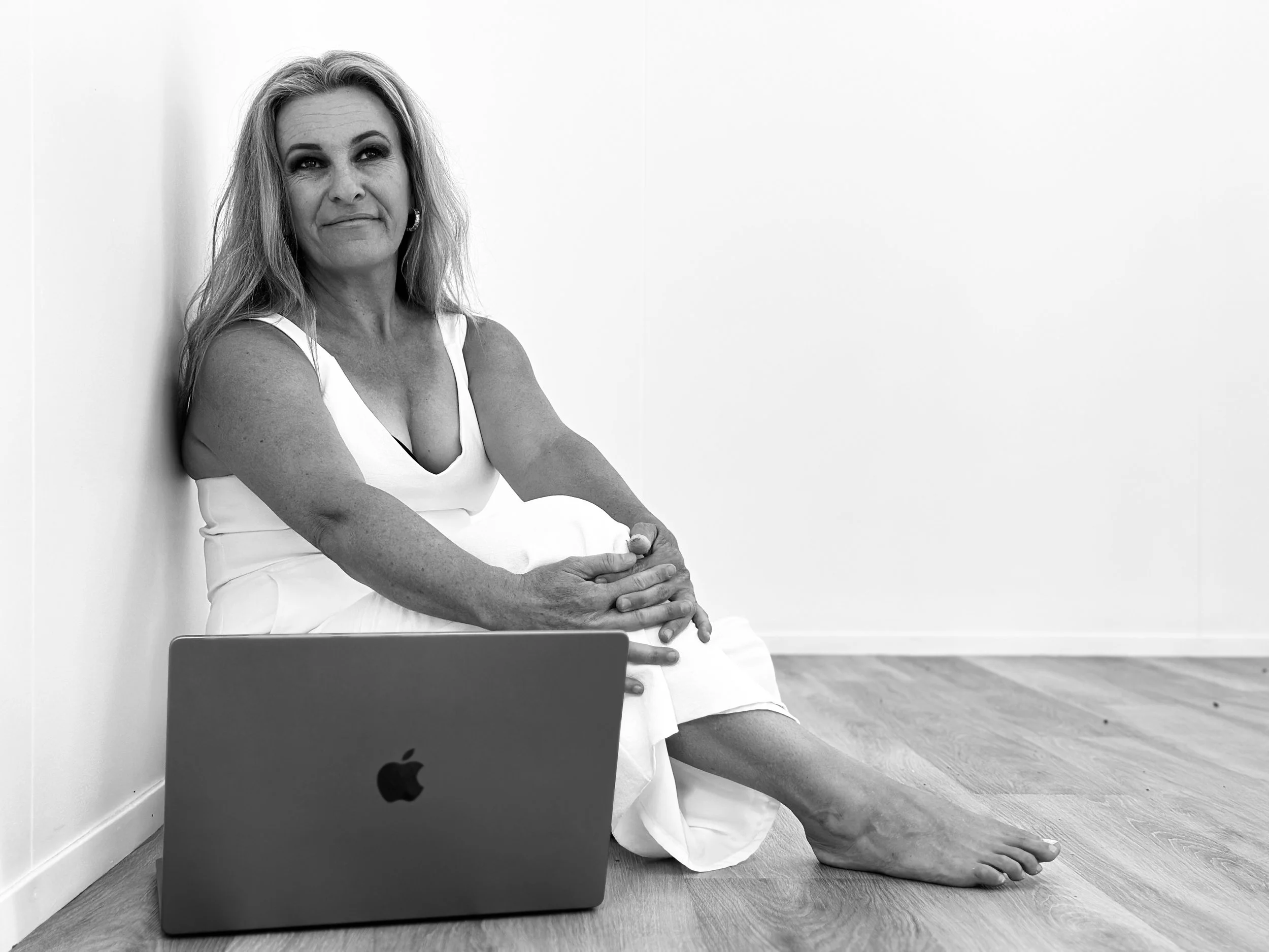 Black and white photo of a woman sitting on the floor with her back against a wall, wearing a white dress, with an open MacBook in front of her.