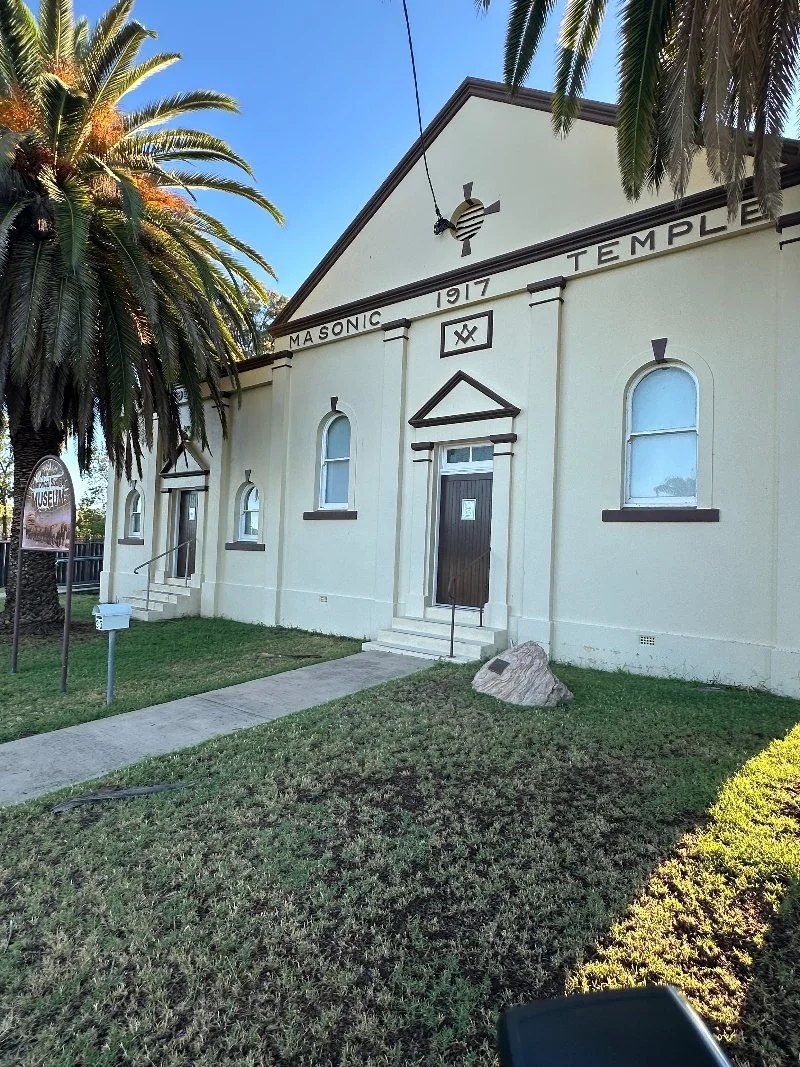 The front of a white masonry building labeled as Masonic Temple, built in 1917, with a triangular pediment, arched windows, a staircase leading to a door, and a sign indicating it is a museum. There are palm trees and a grassy lawn surrounding the building.