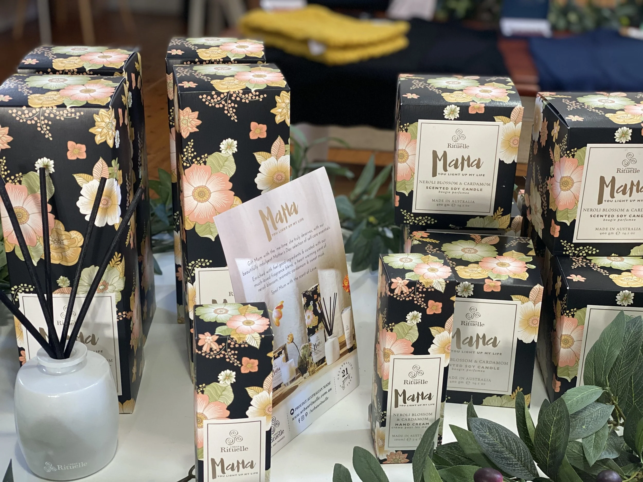 Display of floral-themed boxes and candles with the label "Mama" and a reed diffuser, all featuring a black background with pink, white, and yellow flowers, arranged on a white table with some green leaves.