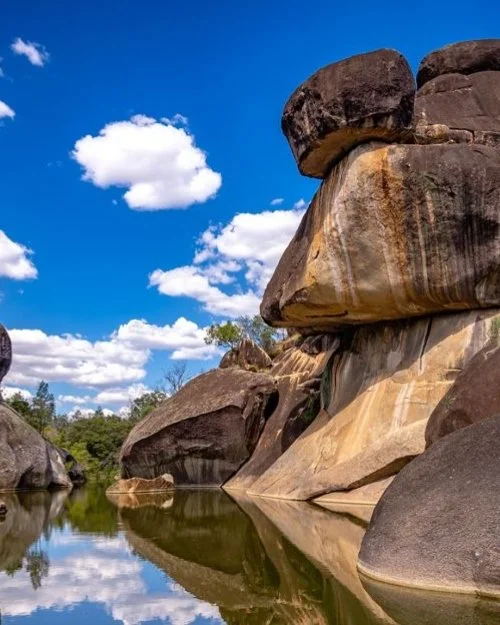 Large rock formations beside a calm river under a blue sky with scattered white clouds.