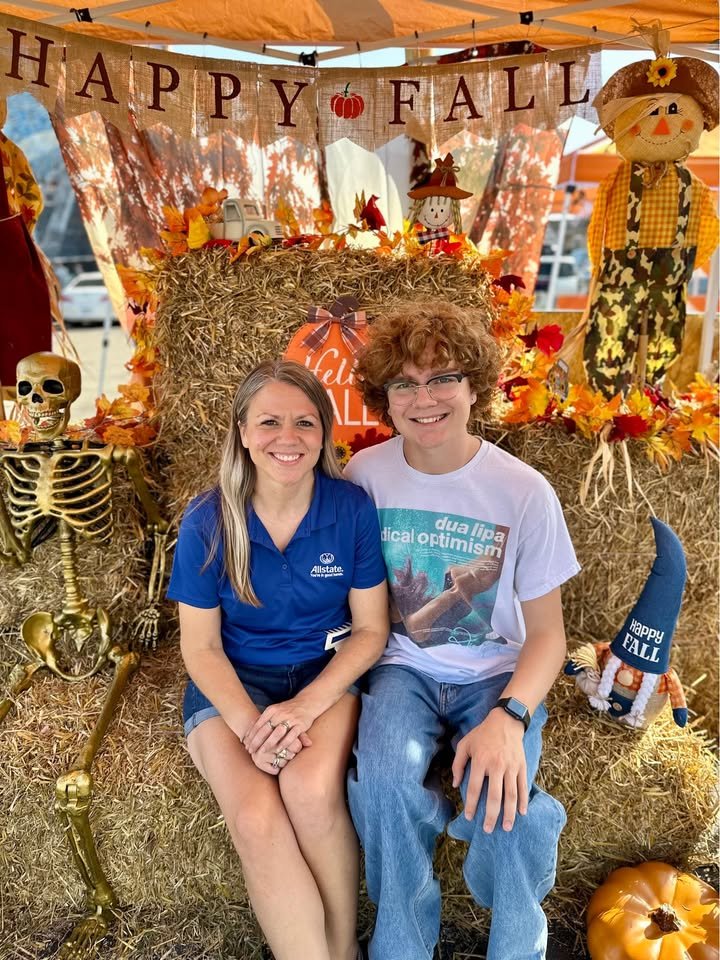 Two people sitting on hay bales in front of fall decorations. The backdrop includes a banner that says "Happy Fall," a scarecrow, pumpkins, leaves, a skeleton, and a stuffed gnome wearing a blue hat with "Happy Fall" on it.