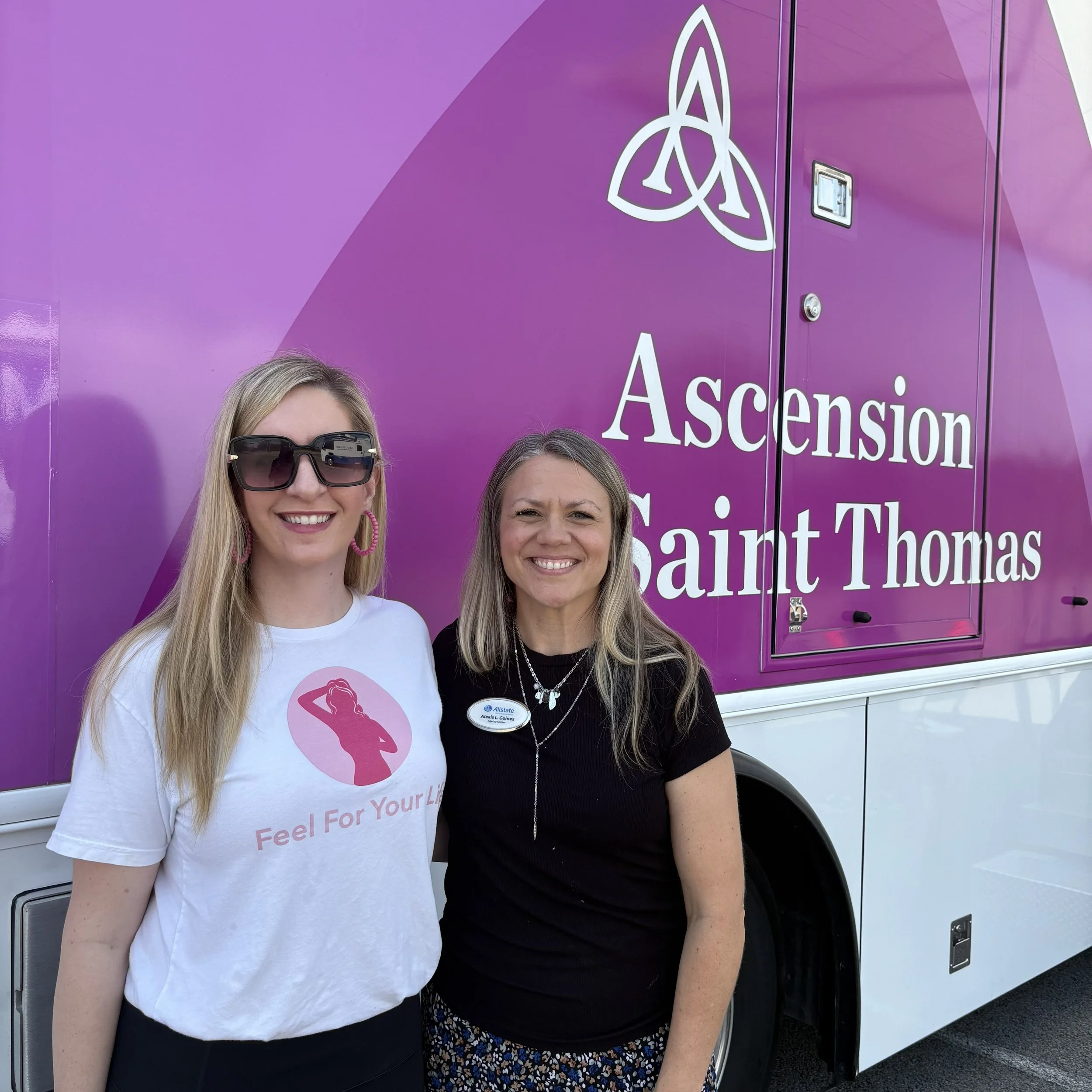 Two women standing in front of a purple mobile health unit with the words "Ascension Saint Thomas" and a logo. One woman is wearing sunglasses and a white t-shirt with a pink silhouette and the phrase "Feel For Your Life." The other woman is smiling, wearing a black shirt and a name tag.