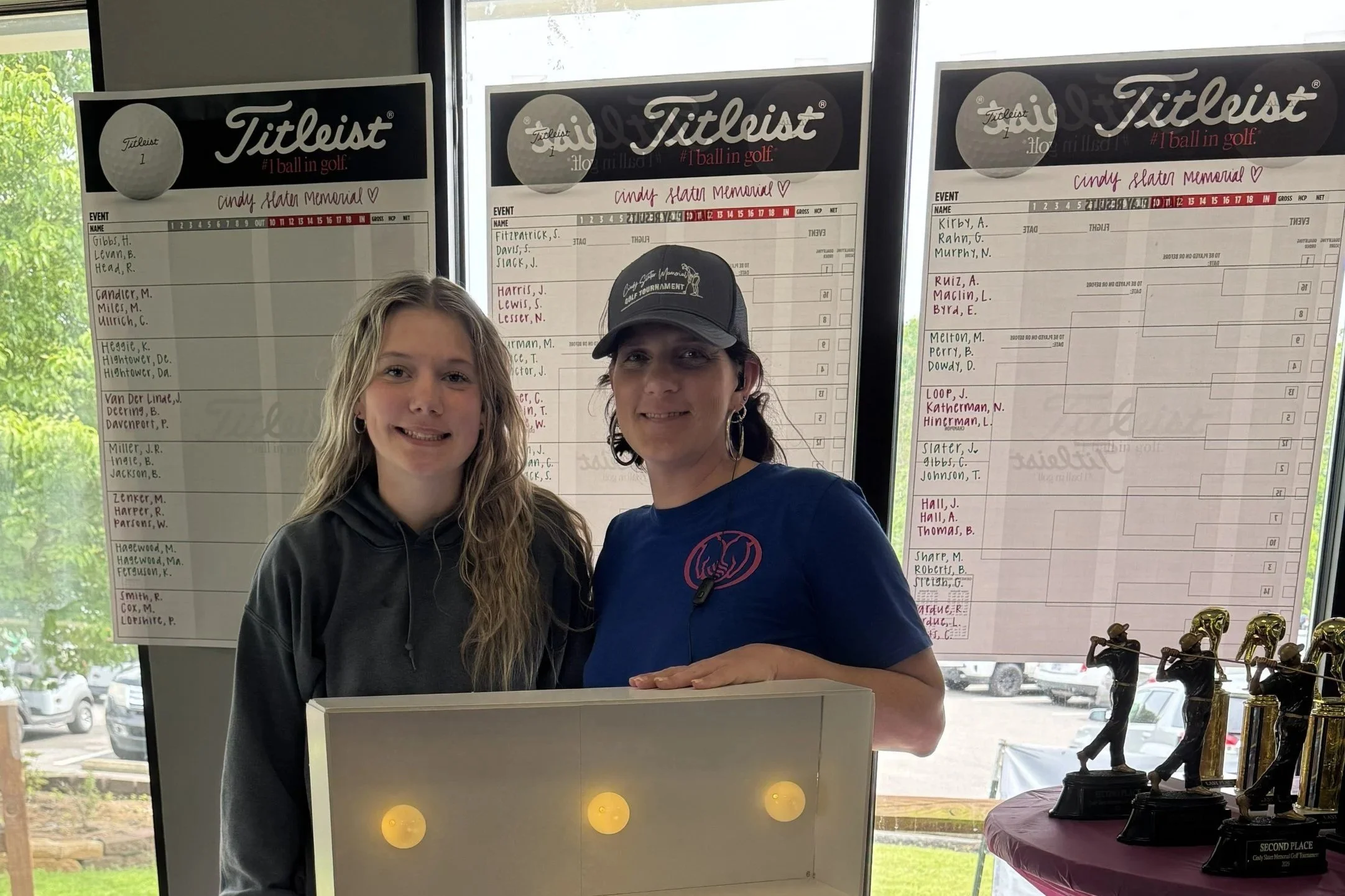 Two young women stand in front of scoreboards at a golf event, smiling. One wears a black hoodie, the other a blue T-shirt, and a baseball cap. Trophies are displayed on a table to the right.