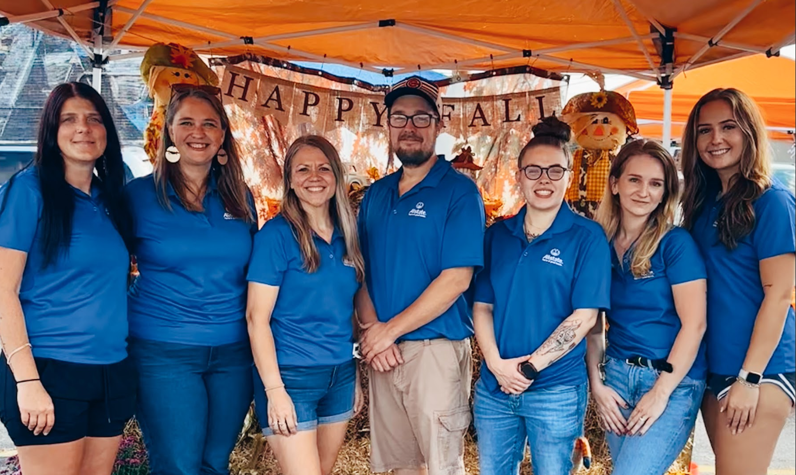 Group of seven people standing together at an outdoor fall festival, wearing matching blue shirts, with a decorated fall themed backdrop and a 
