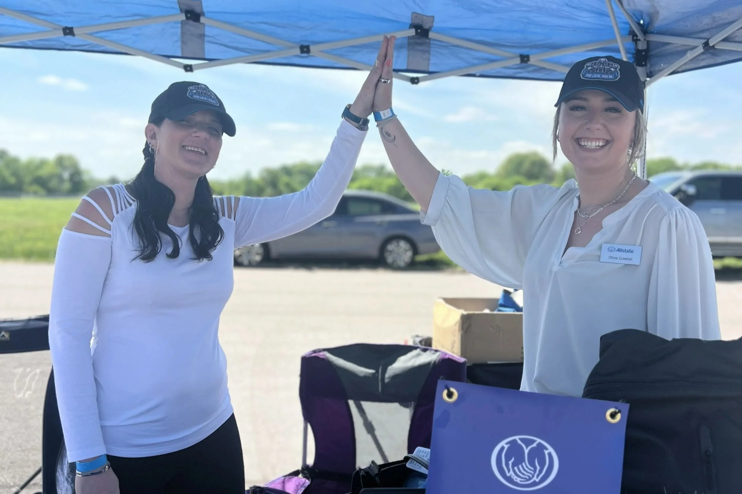 Two women under a blue canopy, giving each other a high five. One woman is wearing a white long sleeve top and a black cap, and the other is wearing a white shirt, a black cap, and a name tag that reads 'Olivia Cuthill'. They are smiling and standing outdoors with cars in the background.