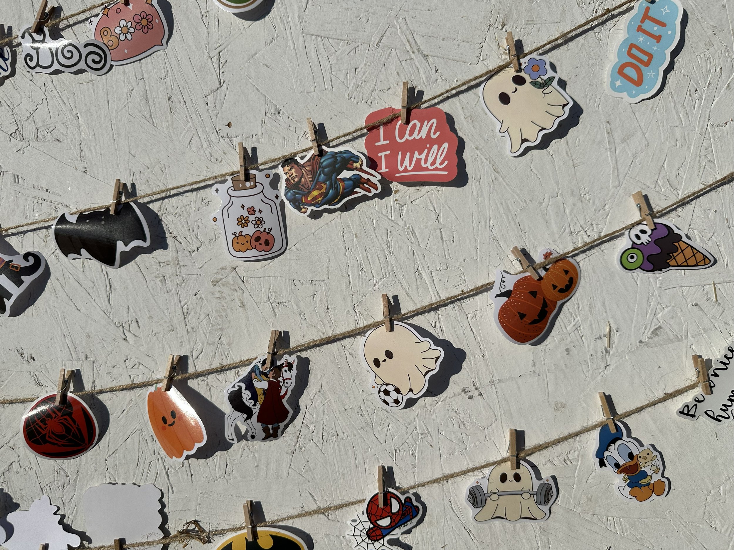 Decorative paper cutouts with Halloween and cartoon themes hanging on strings with clothespins. The cutouts include a ghost, pumpkin, spider, Batman, Spider-Man, Superman, Disney characters, and motivational messages.