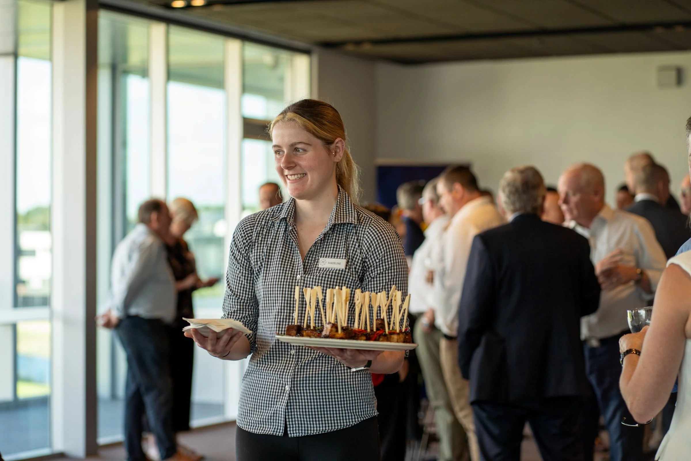 Modern conference room with round tables and chairs, large windows, and a few people sitting and standing.