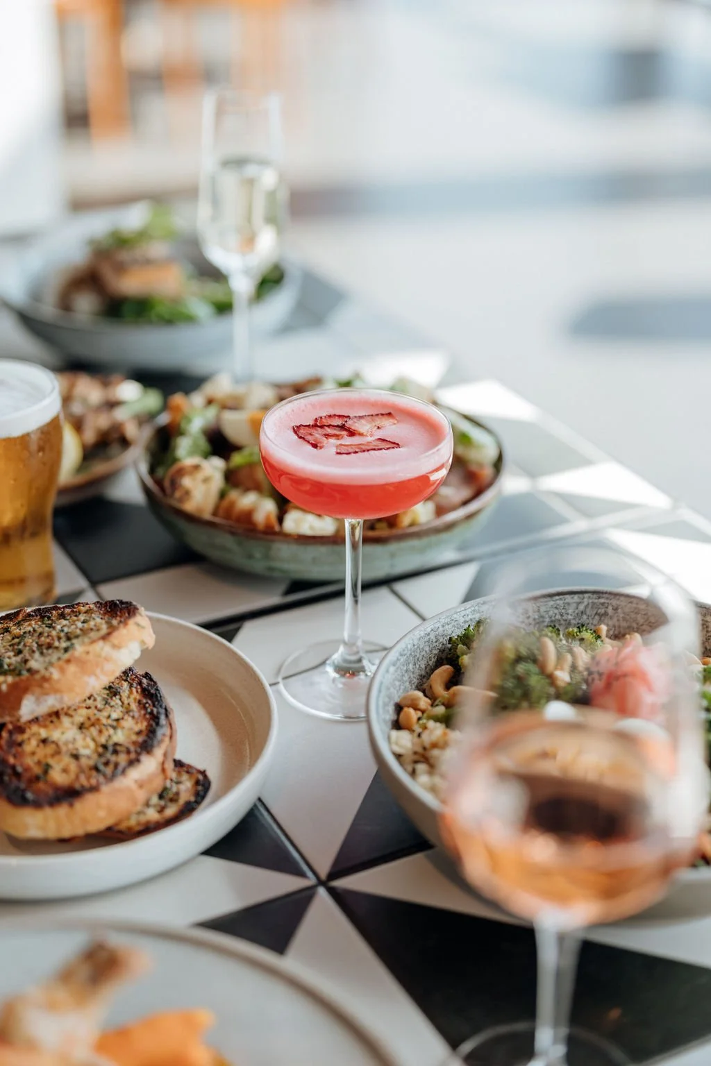 A table with food and drinks including a pink cocktail with ice, a beer, salad, and grilled bread in a restaurant setting.