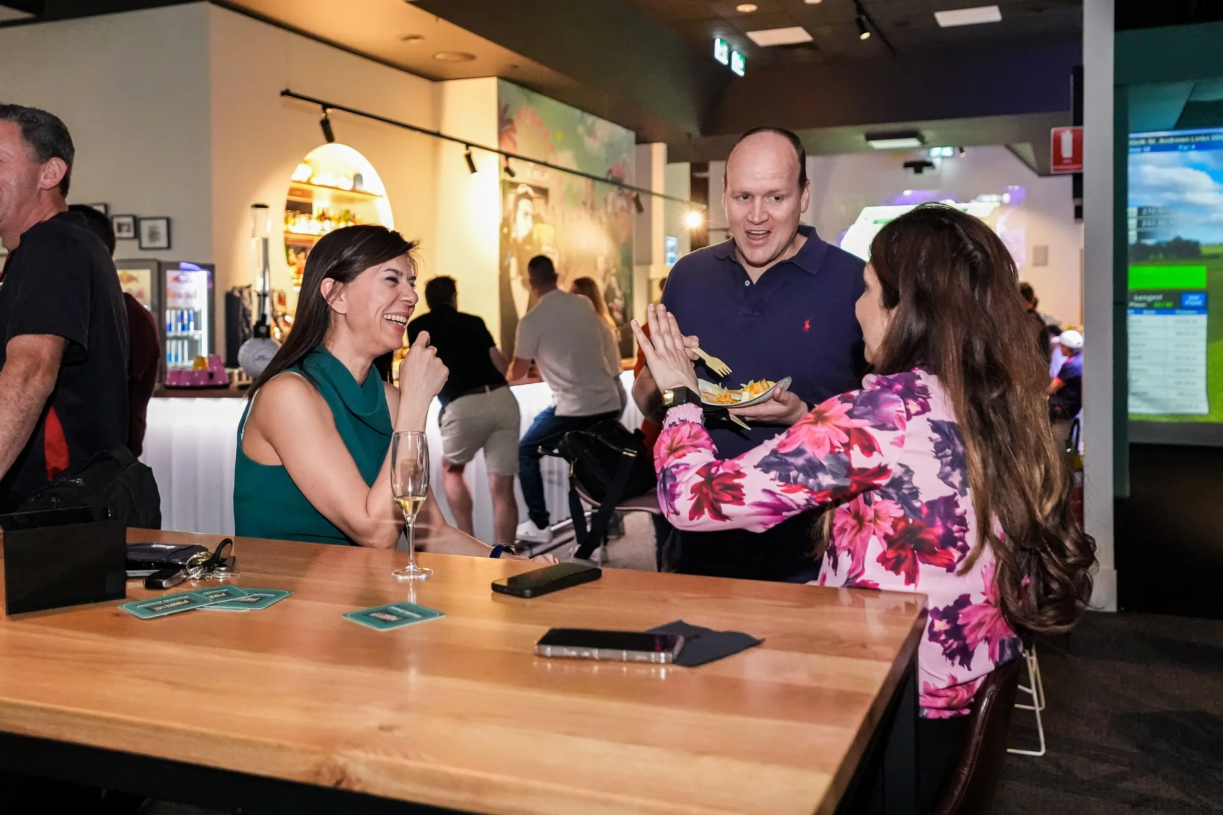 A group of three people, two women and a man, are cheerful and laughing at a bar or restaurant. The woman on the left has long dark hair and is wearing a green sleeveless top, while the woman on the right has long brown hair and is wearing a pink and