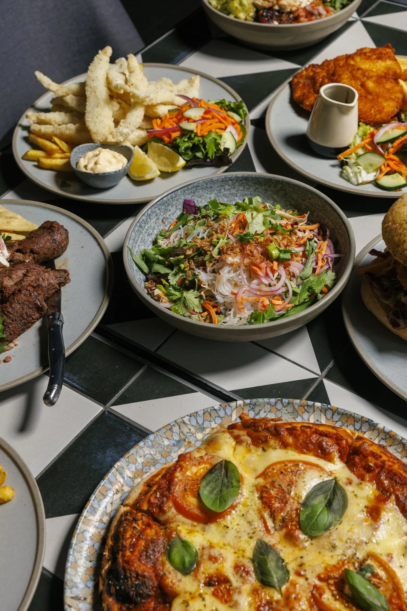 Various plates of food on a table, including a pizza with basil, a bowl of salad, fried calamari with lemon wedges, fried fish with sauce, and a meat dish.